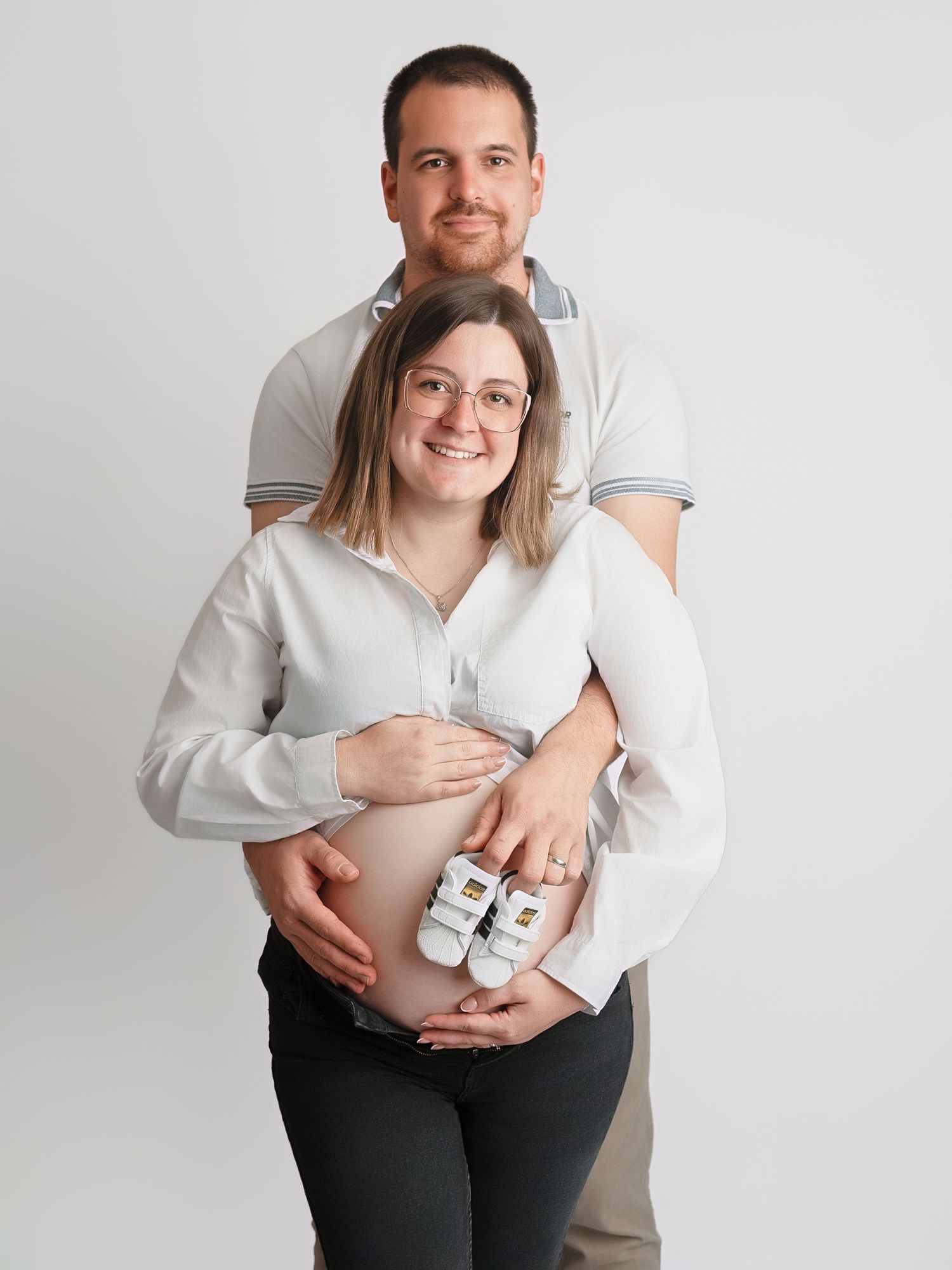 Smiling pregnant couple posing together, woman holding tiny baby shoes against her baby bump in a bright studio setting.