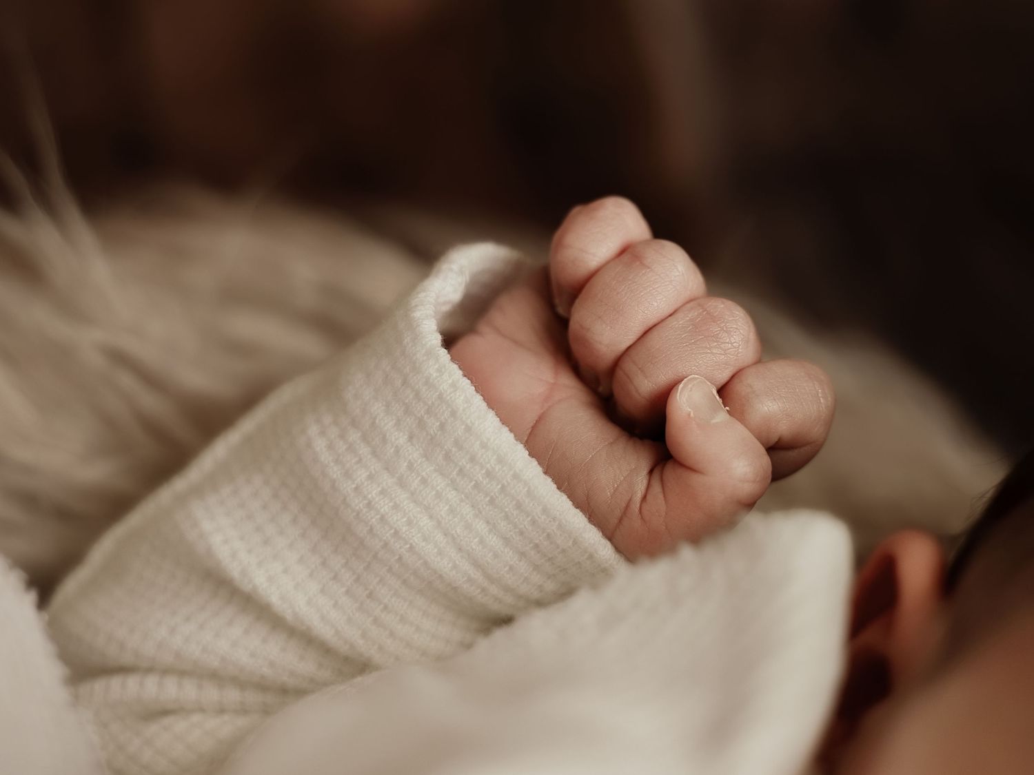 Close-up of a newborn baby's tiny clenched fist in a white knit outfit, capturing delicate infant details.