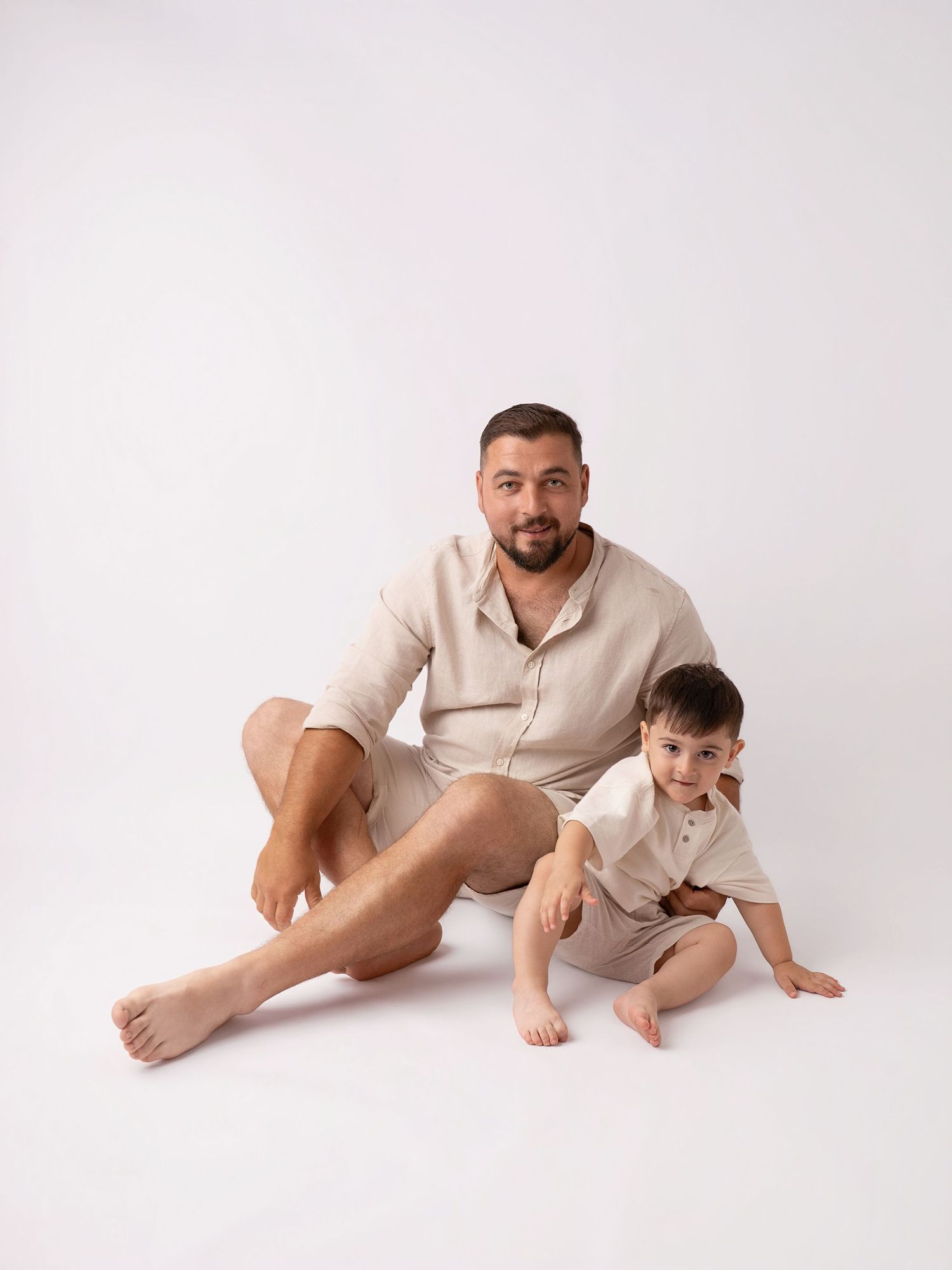 Man and toddler boy in matching beige linen outfits sitting together on white background.