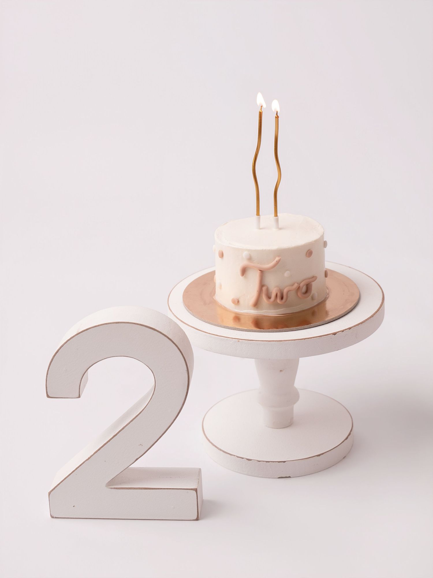 A small birthday cake with two candles on a white cake stand next to a wooden number 2.