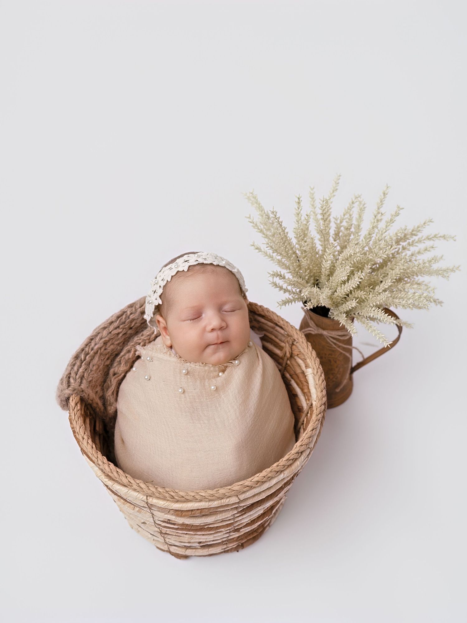 Sleeping newborn baby wrapped in beige swaddle with pearl details, wearing lace headband, posed in wicker basket with dried plant.