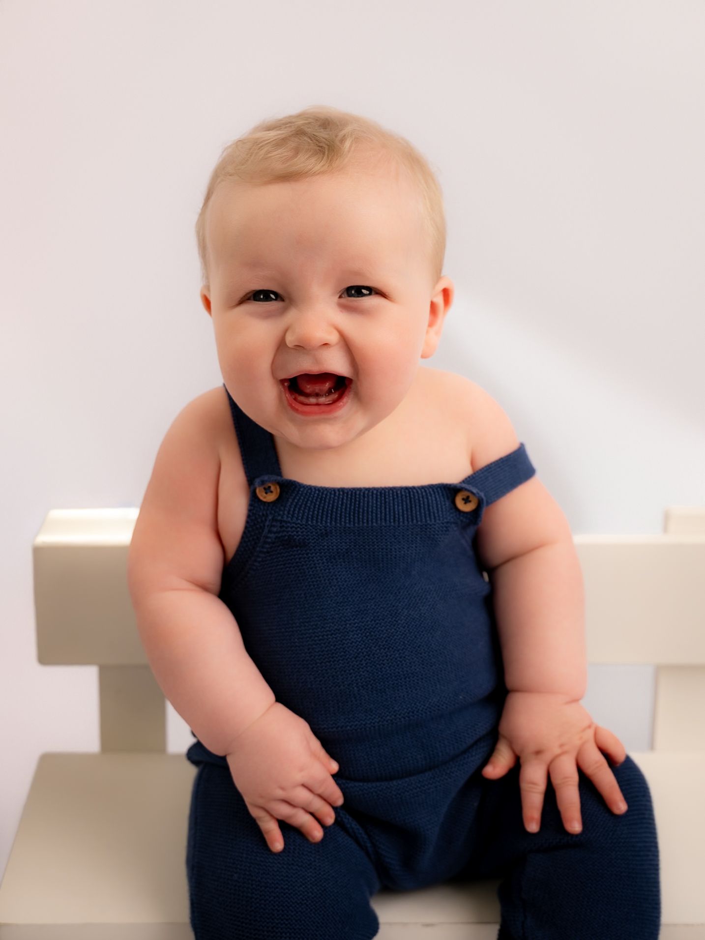 Happy smiling baby wearing a navy knit overalls sitting on a white chair, laughing joyfully.