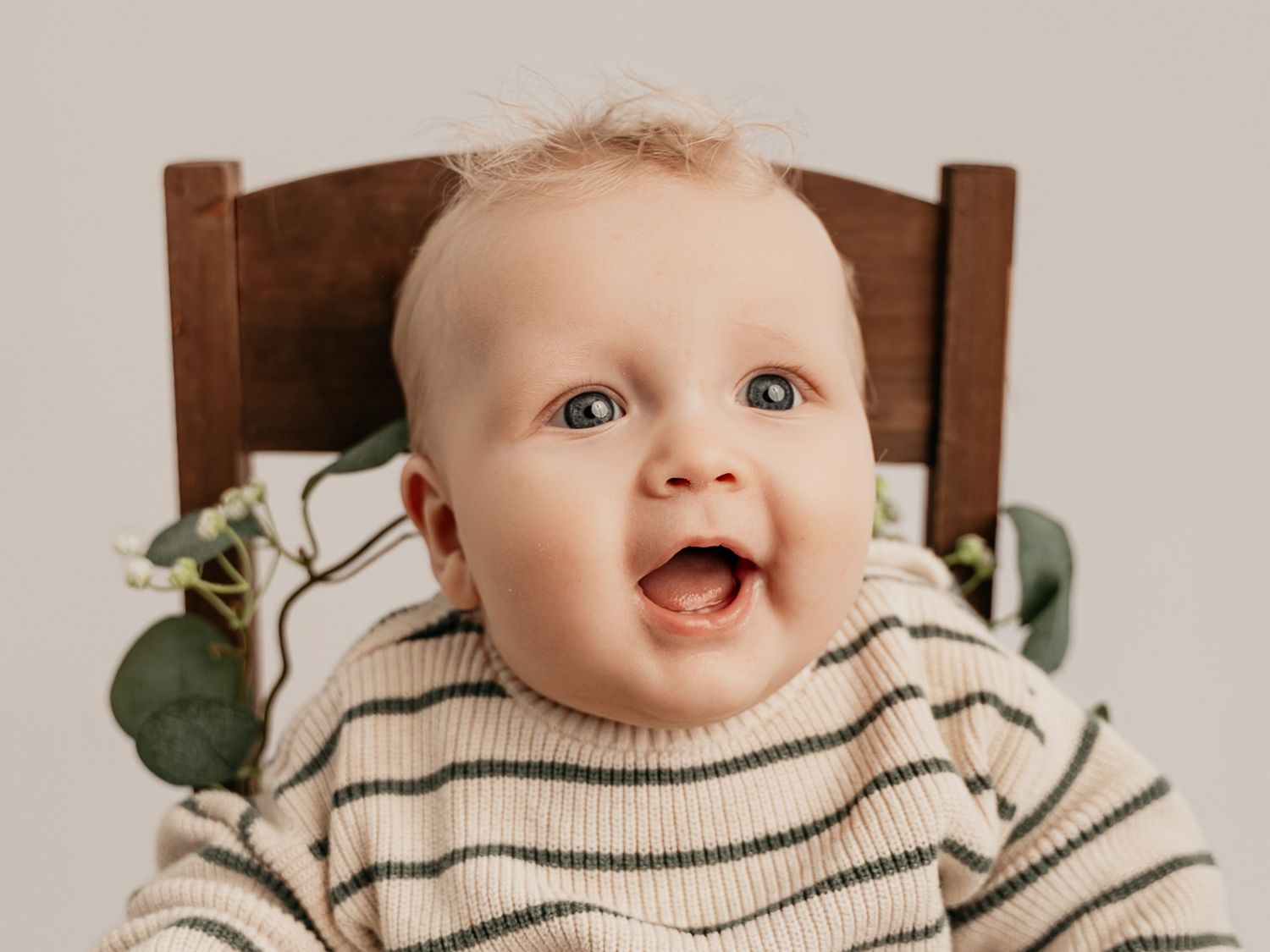 Expressive baby in a striped sweater sitting in a wooden chair with eucalyptus greenery decoration.