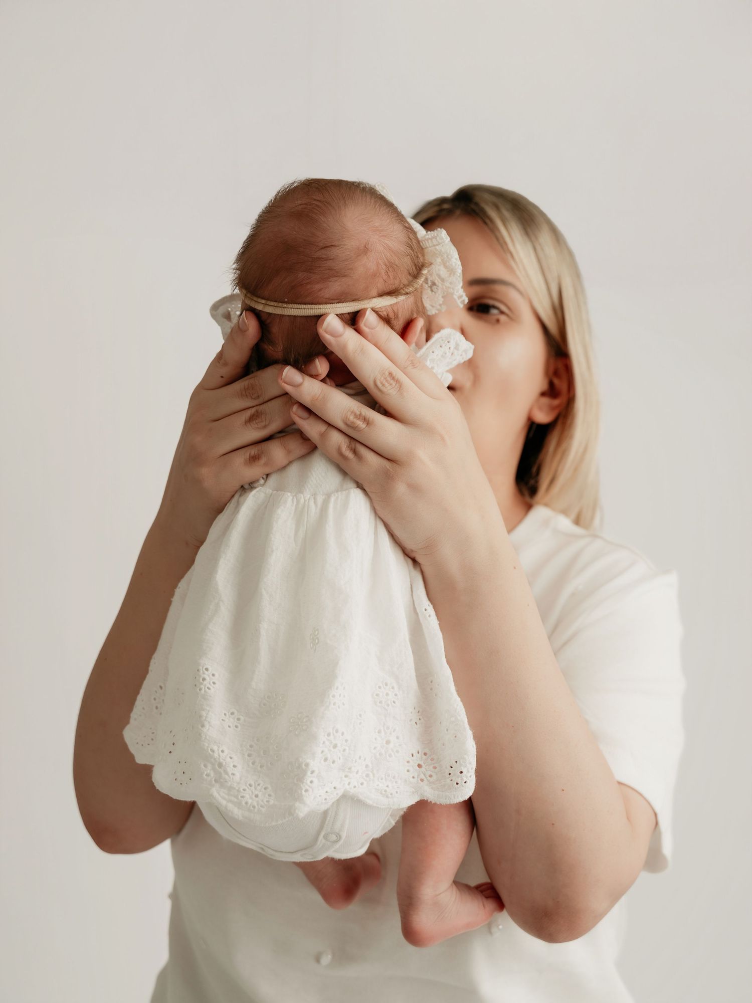 Blonde mother in white shirt kissing newborn baby dressed in white lace outfit against light background.