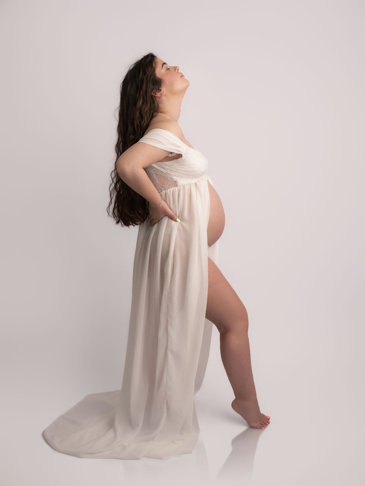 Pregnant woman in a flowing ivory off-shoulder maternity gown posing with head tilted back in a white studio.