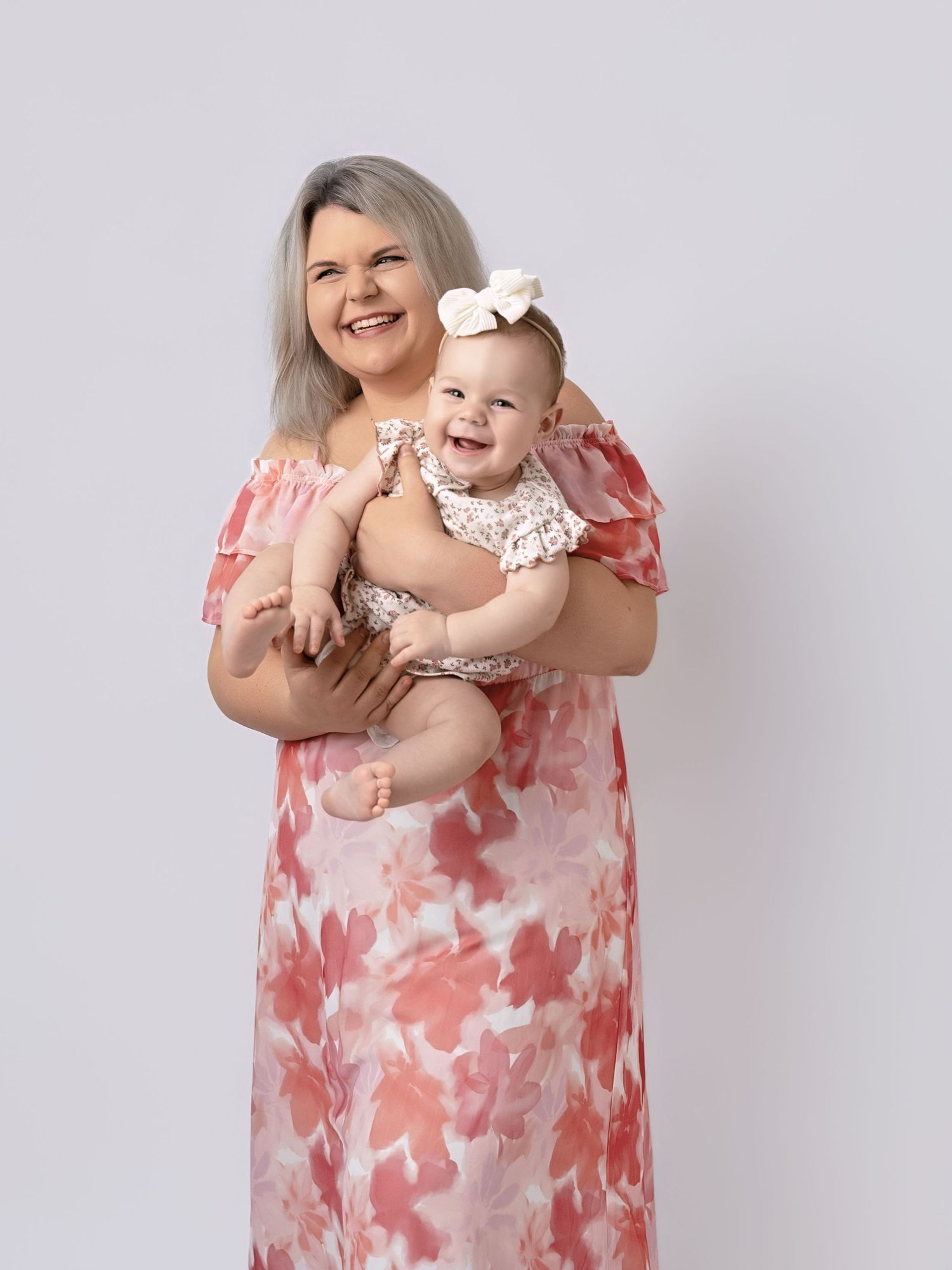 Smiling woman in floral maxi dress holding a happy baby girl with a bow in a studio portrait.