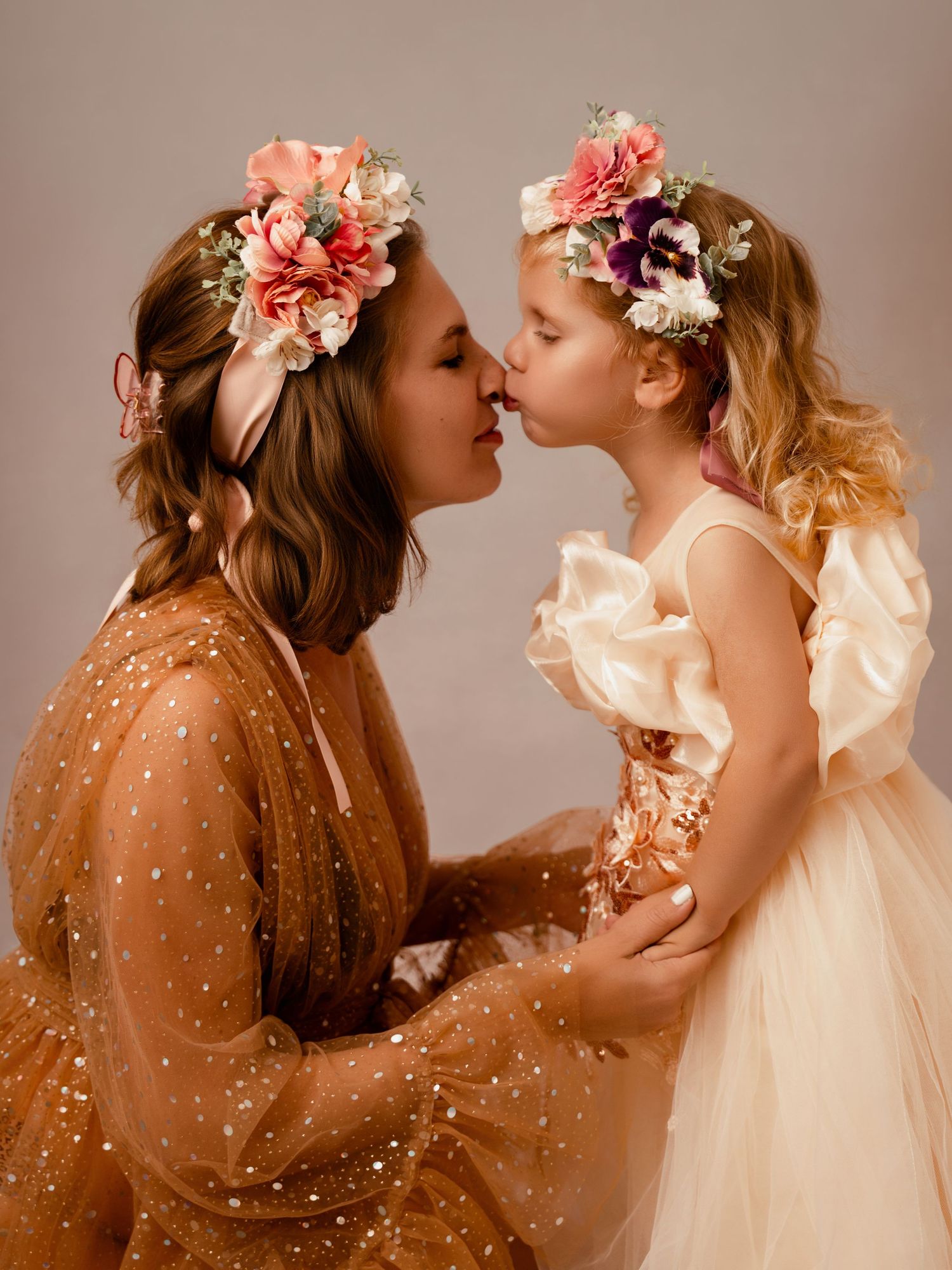 Mother and daughter in matching floral crowns and tulle dresses sharing a sweet kiss in a studio portrait.