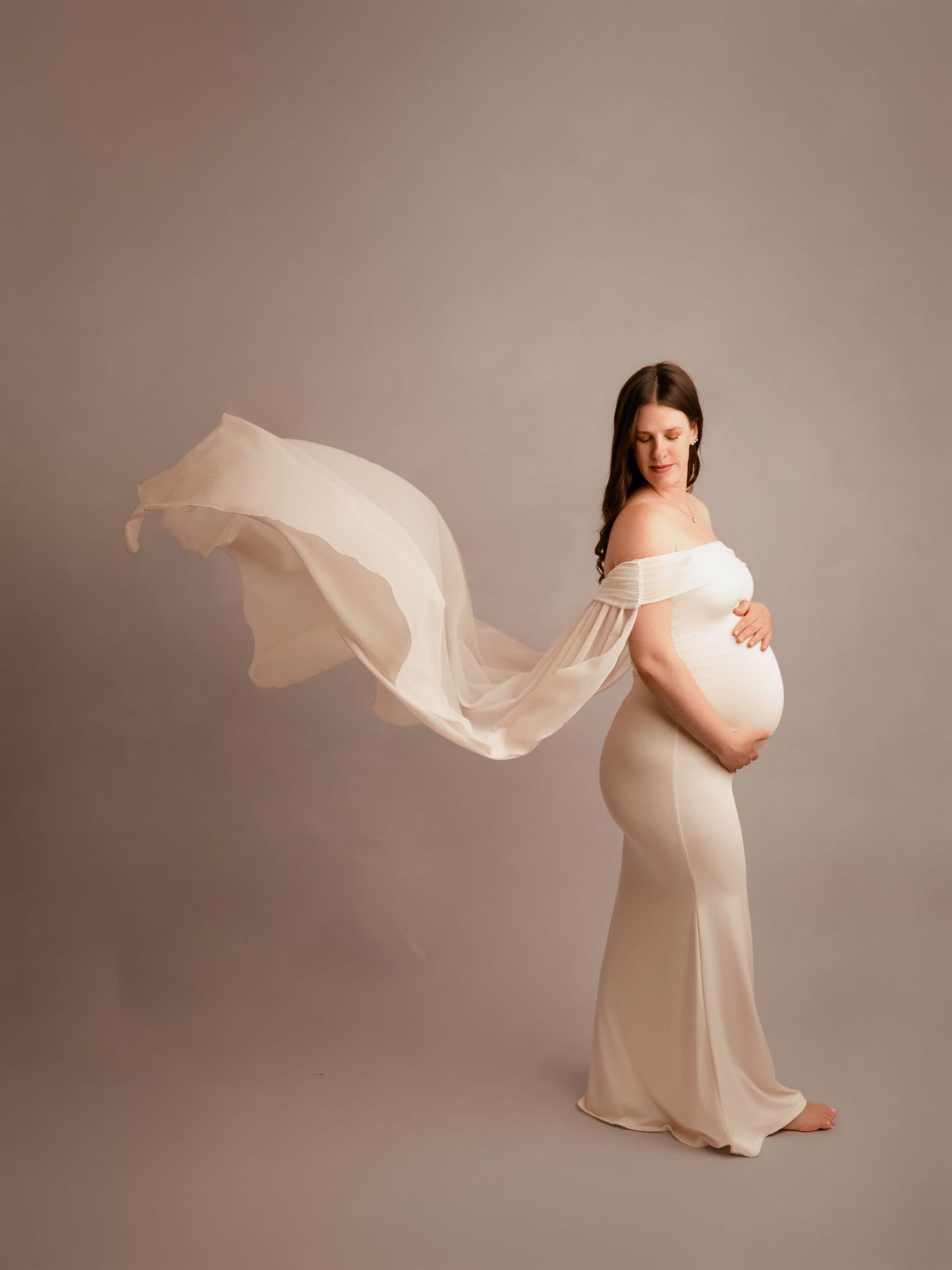 Pregnant woman in flowing white off-shoulder gown with fabric billowing, cradling baby bump in studio maternity portrait.