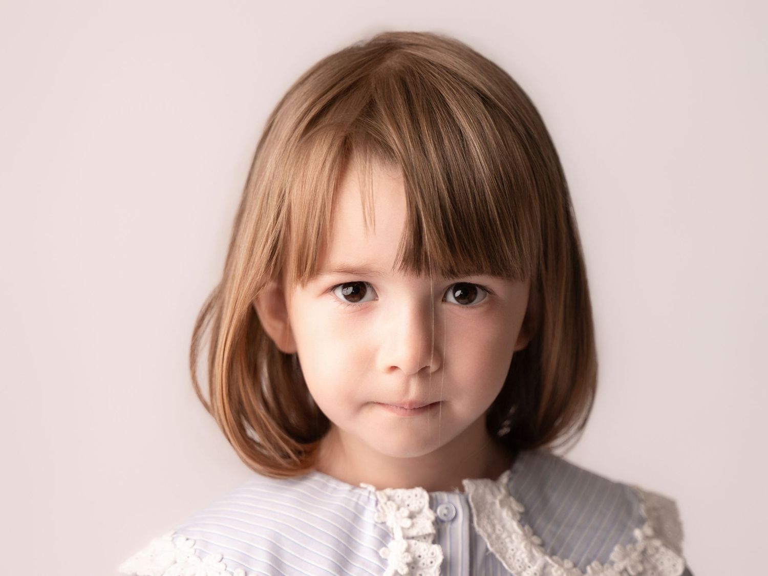 A young girl with a blonde bob haircut wearing a white lace-trimmed dress poses for a studio portrait.