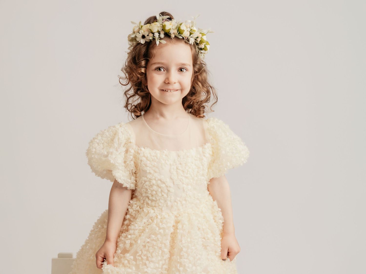 A young girl in a cream textured puff-sleeve dress wearing a delicate floral crown smiles softly.