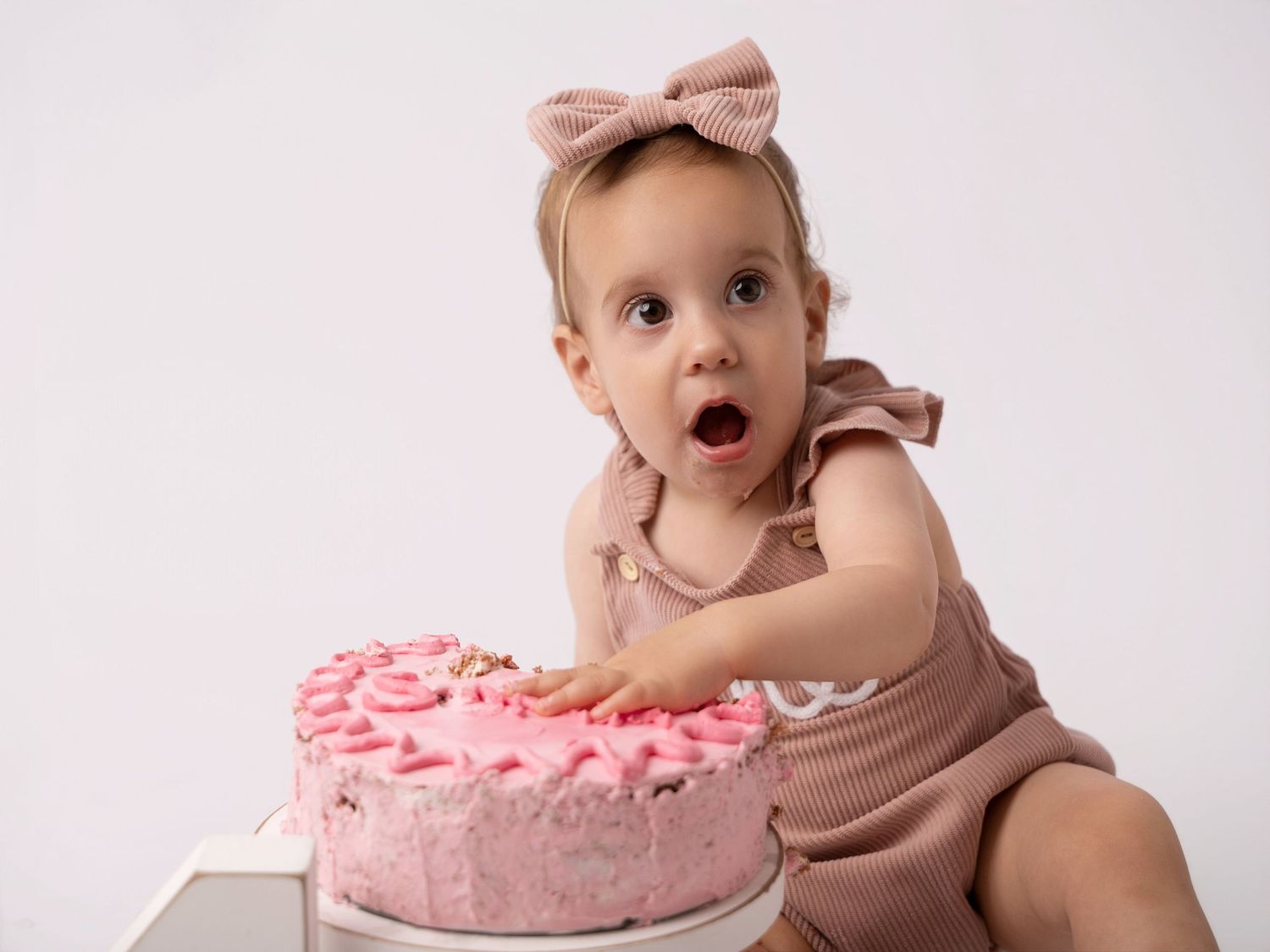 A surprised baby girl in pink romper reaches toward a heart-shaped pink birthday cake.