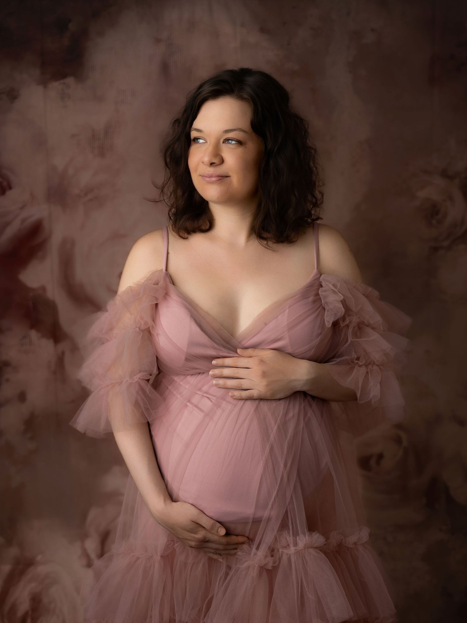 Pregnant woman in pink tulle off-shoulder maternity gown poses gracefully against a floral painted studio backdrop.