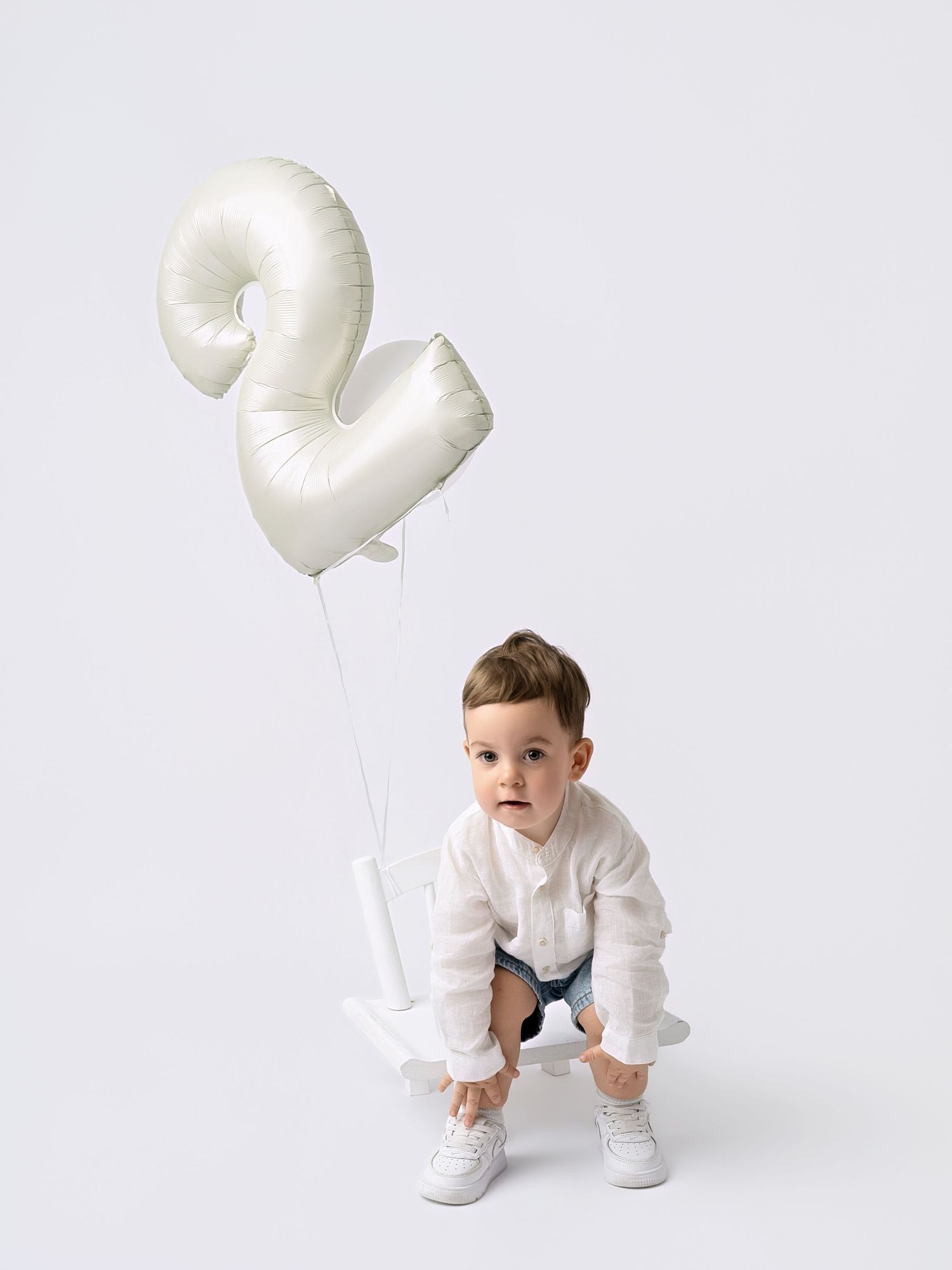 Toddler boy in white shirt and denim shorts crouching beside white number 2 balloons on white background.