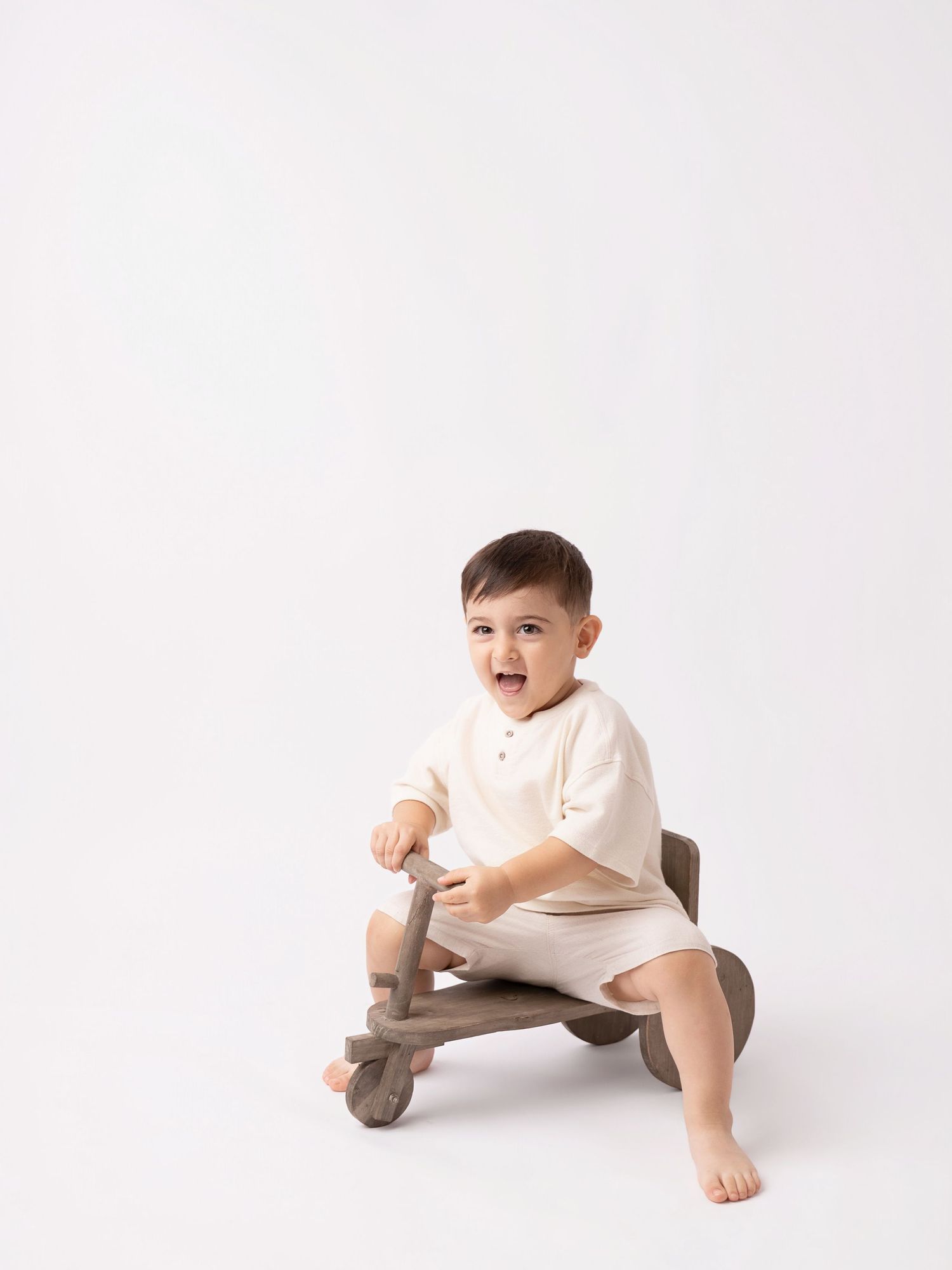 Smiling toddler boy in cream romper sitting on a wooden balance bike toy against white background.