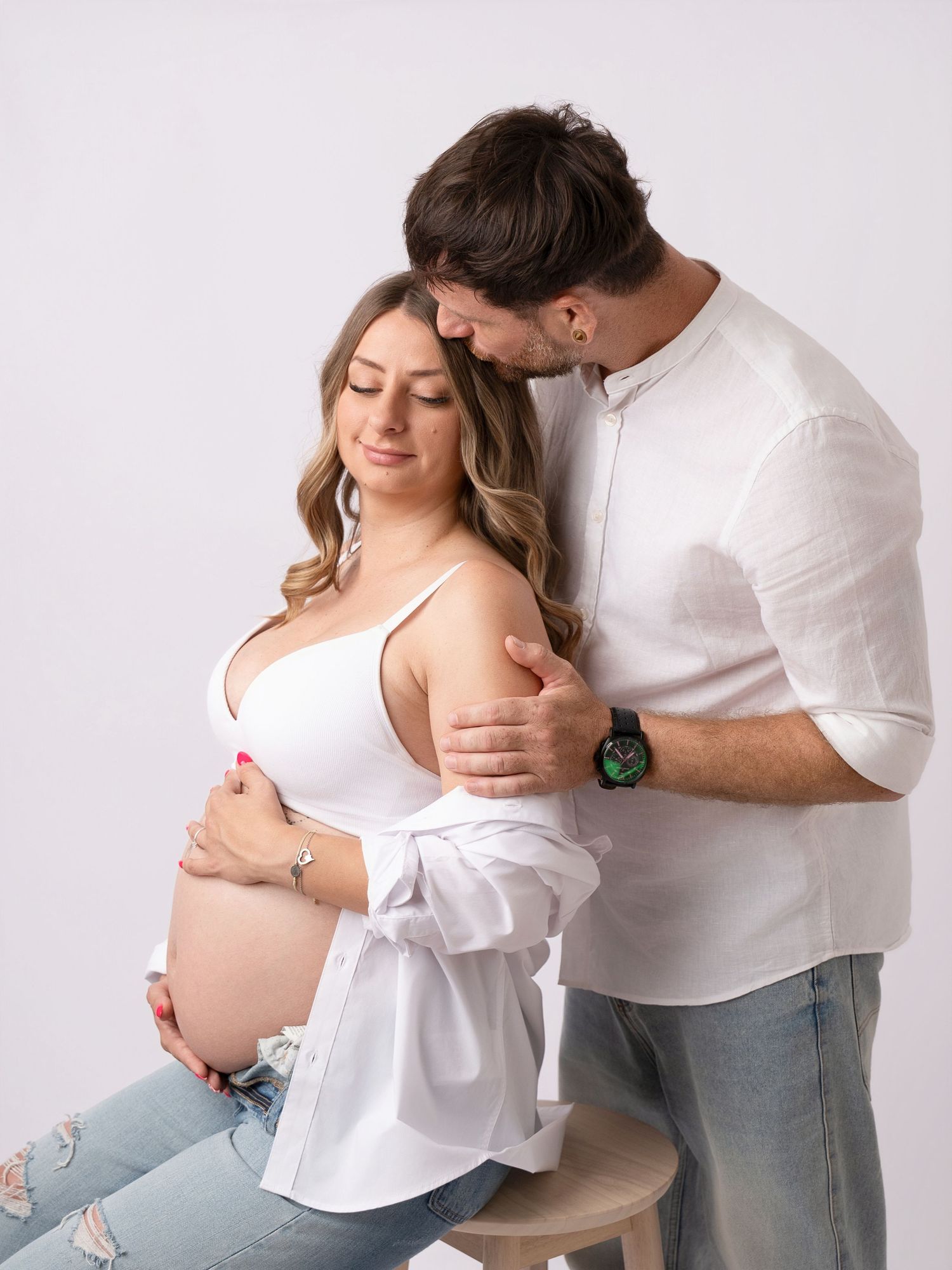 Couple in white outfits posing for maternity photo shoot, man embracing pregnant woman in studio.