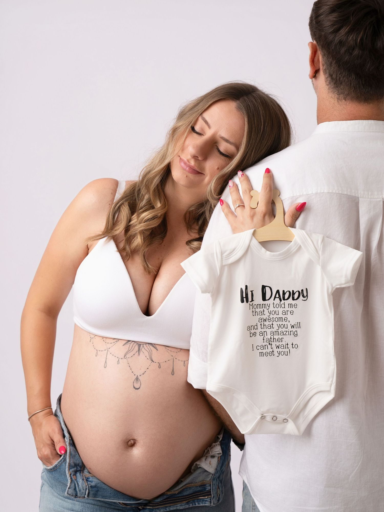 Pregnant woman in white bralette holds baby onesie with partner during maternity photoshoot.