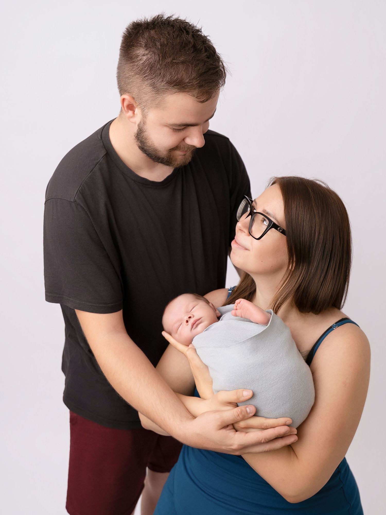 Young couple smiling lovingly at each other while holding their swaddled newborn baby together.