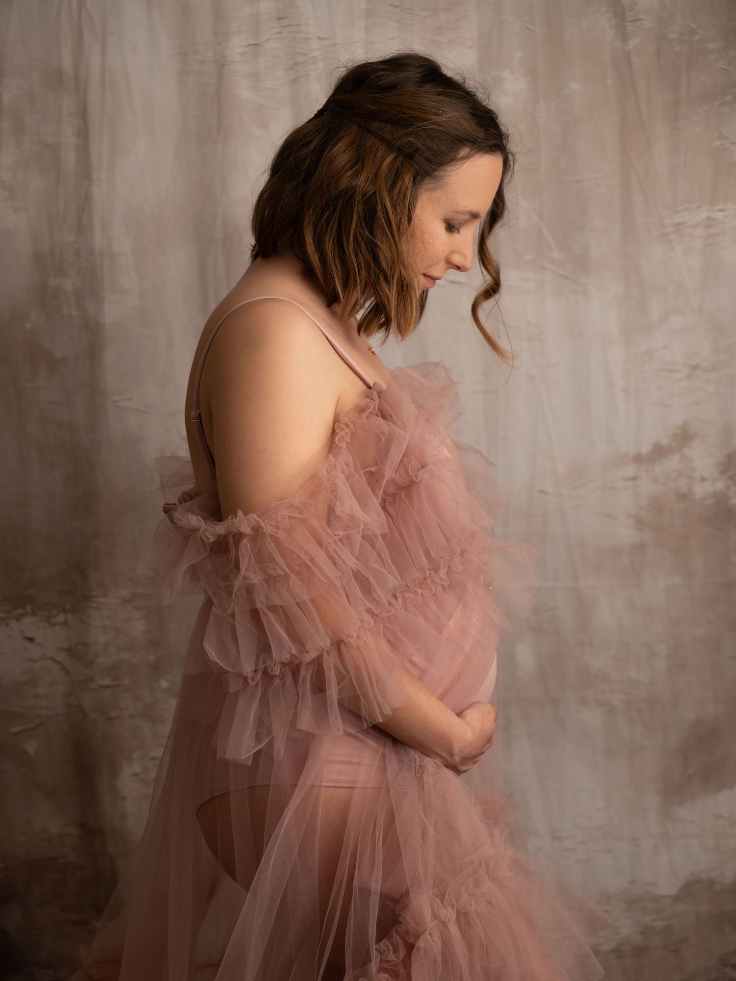 Pregnant woman in pink tulle maternity gown cradling baby bump in professional studio portrait.