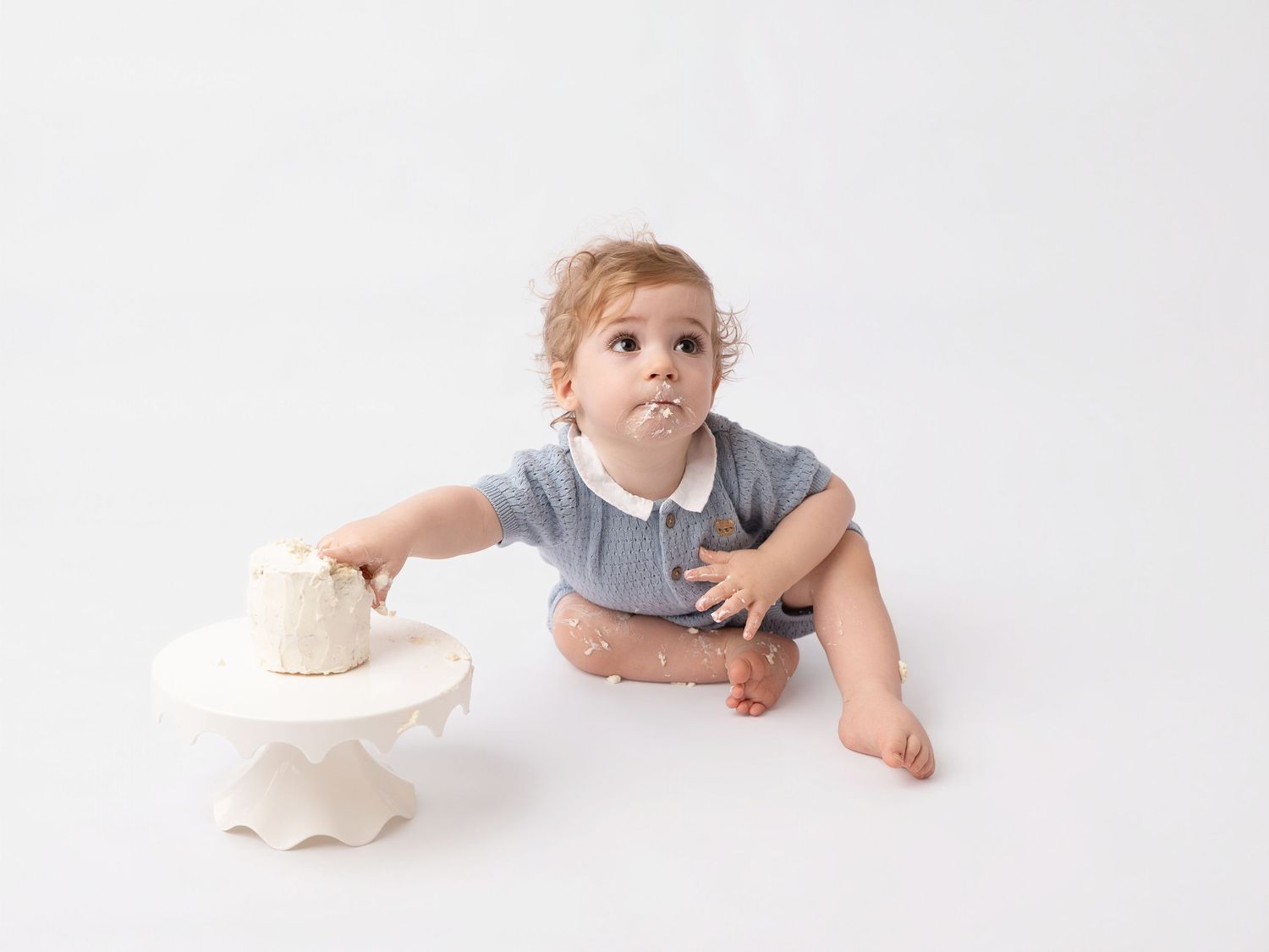 Baby in gray romper touching a white smash cake on a pedestal stand against a white background.