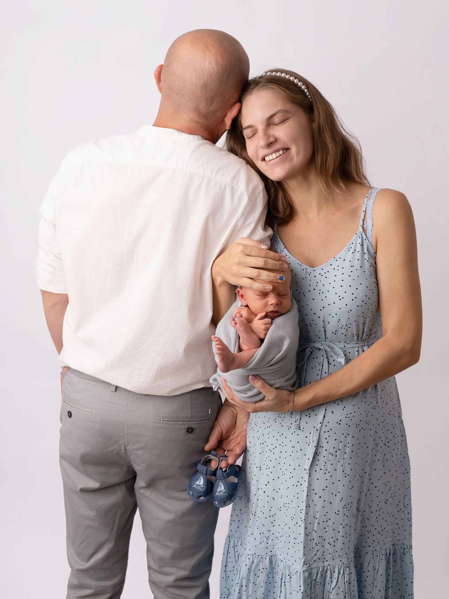 A smiling mother in a blue dress holds her newborn baby while a bald father in white shirt leans in tenderly.