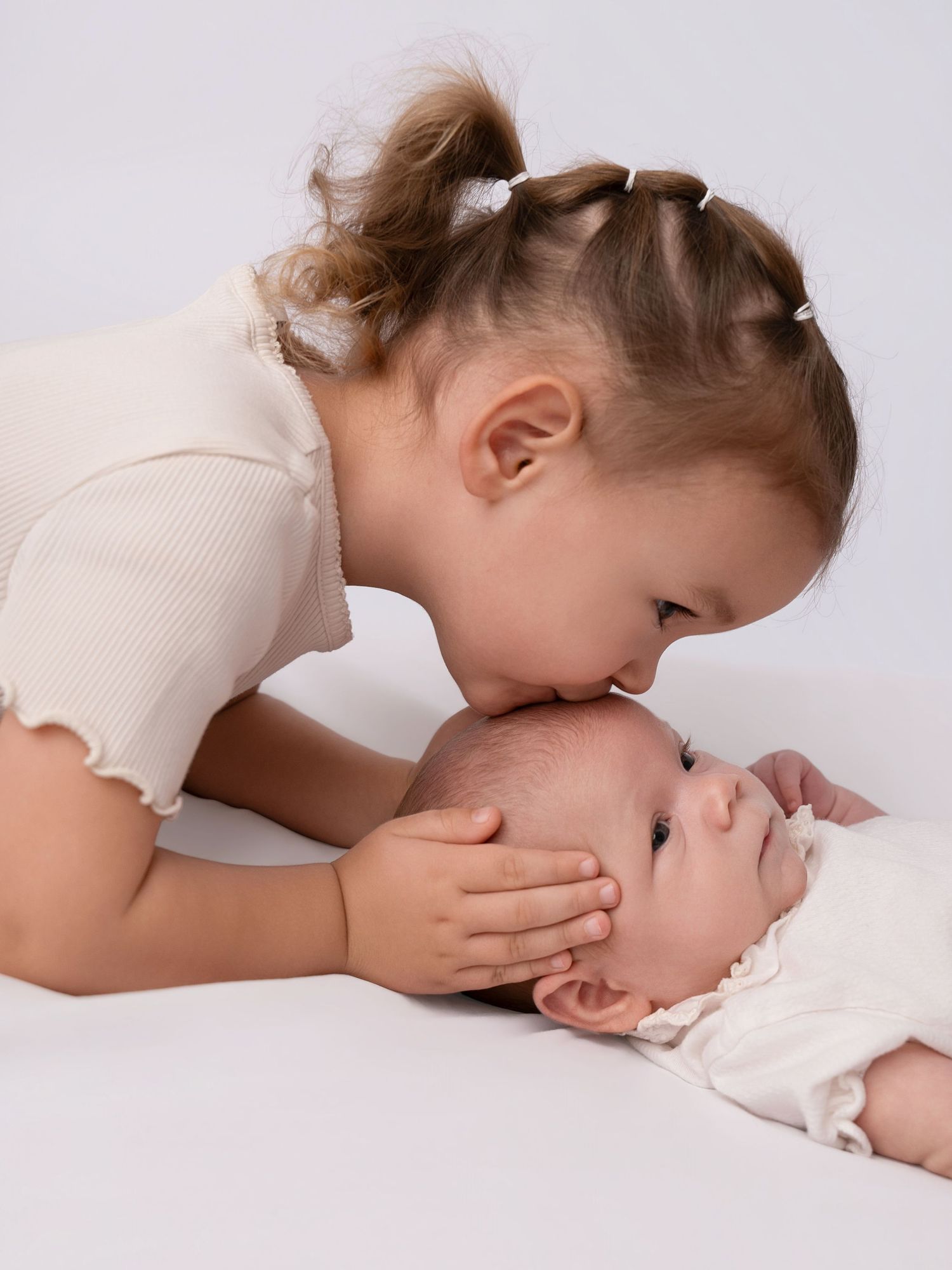 Young girl gently kisses newborn baby on the forehead in a sweet sibling moment on white background.