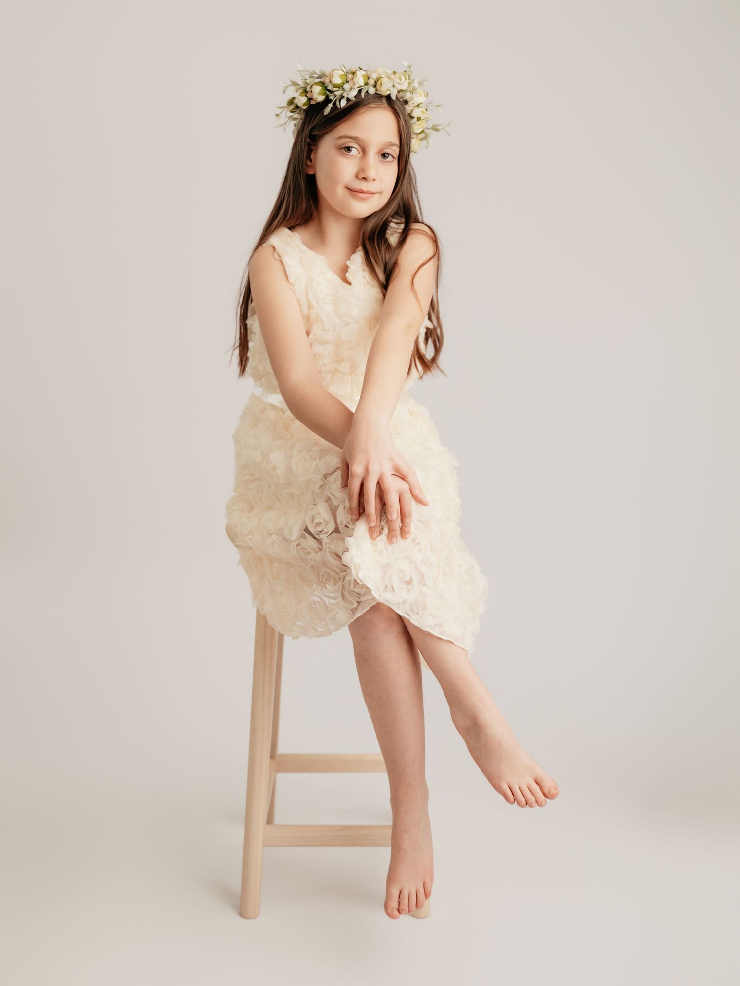 Young girl in cream floral dress and white flower crown sitting on wooden stool in studio.
