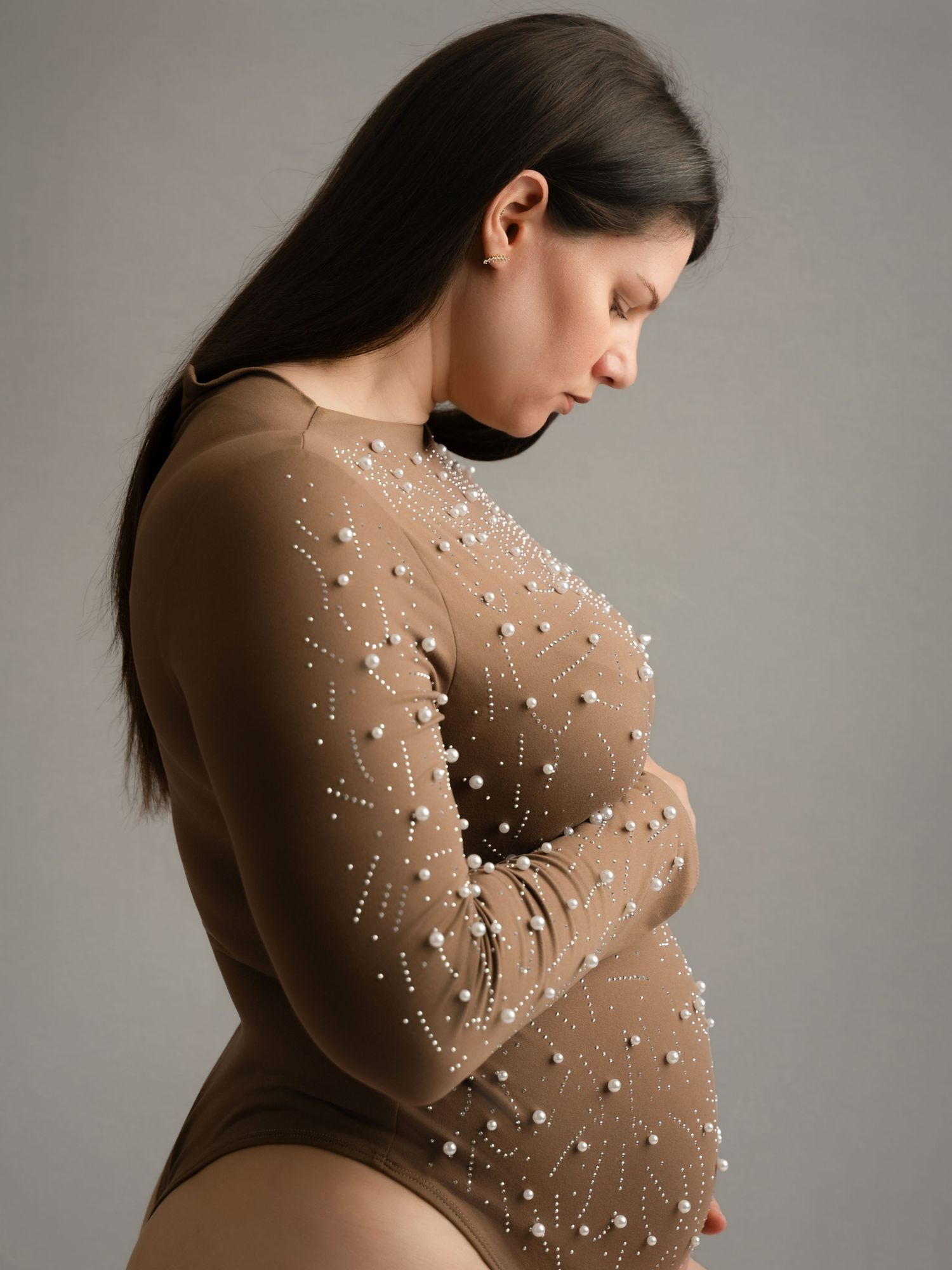 Pregnant woman in brown pearl-embellished bodysuit cradling baby bump in studio portrait.
