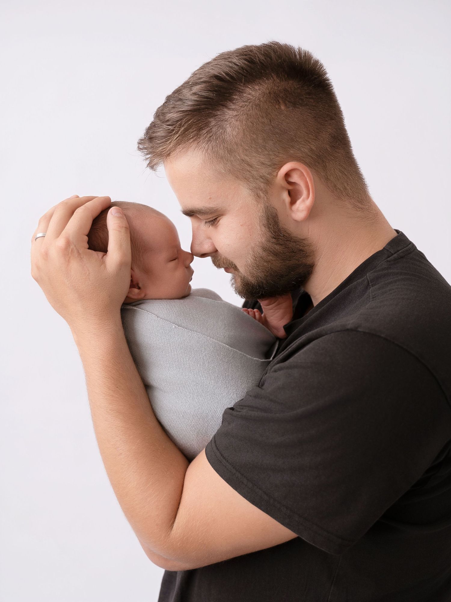 A bearded father tenderly holds his swaddled newborn baby close, touching foreheads in a loving moment.