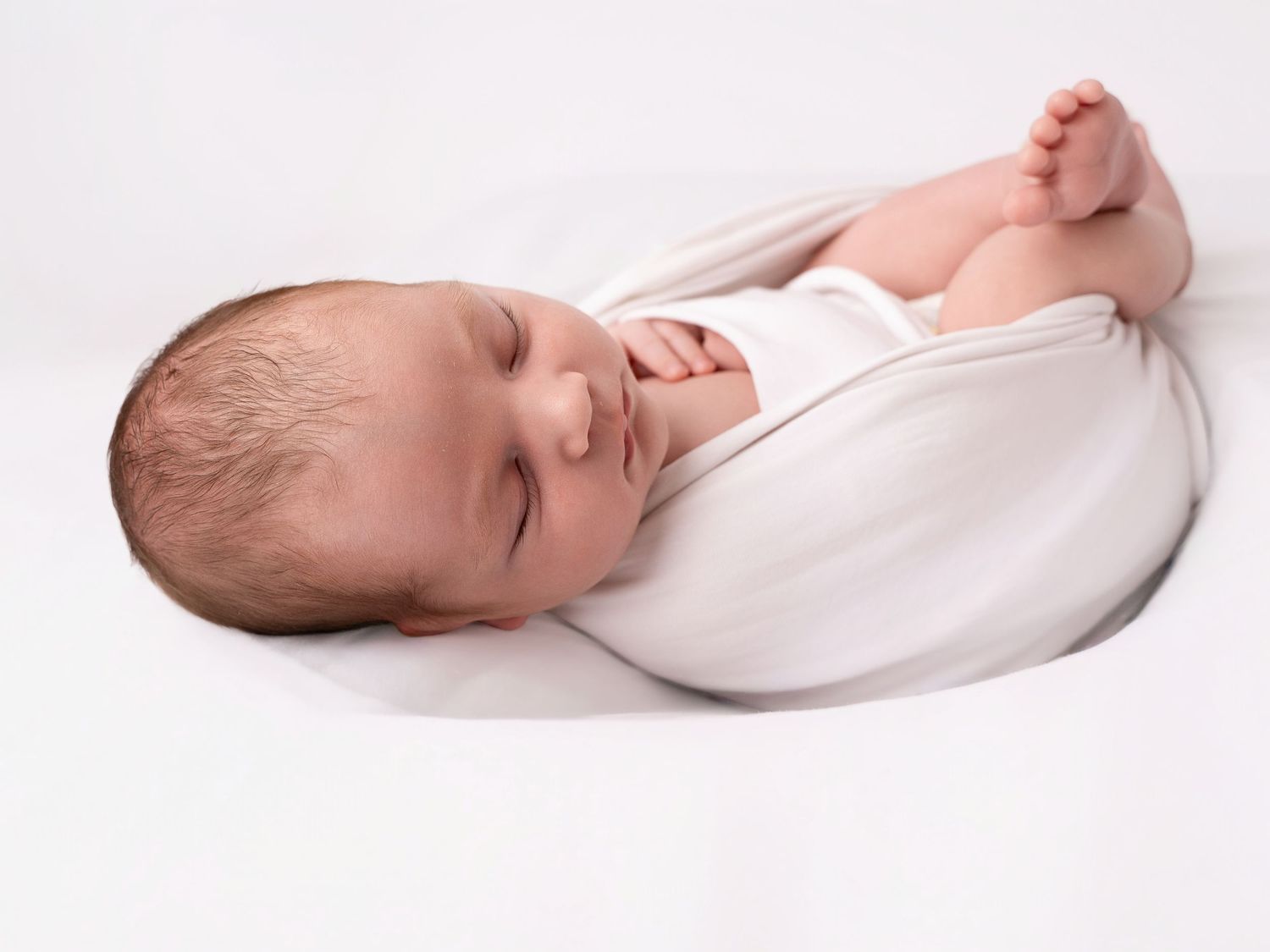 Newborn baby sleeping peacefully swaddled in a white blanket on a soft white surface.