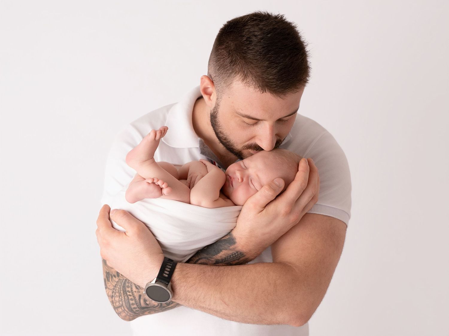 A tattooed father tenderly kisses his sleeping newborn baby while cradling them in his arms against a white background.