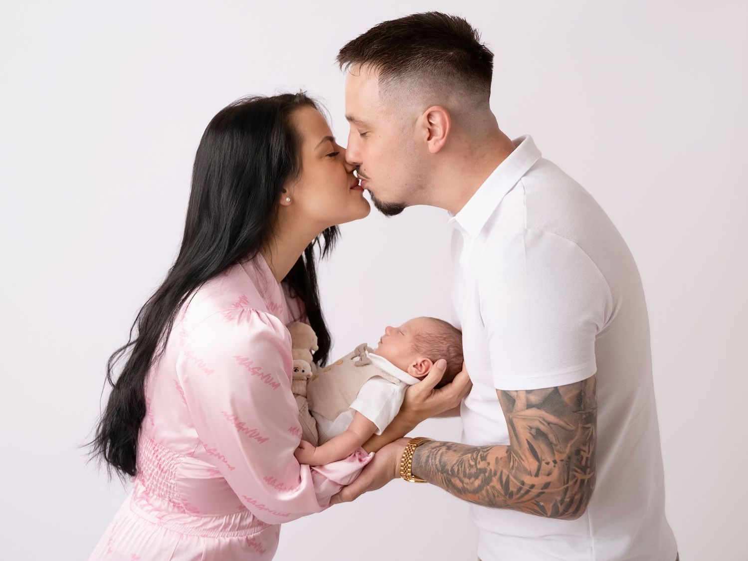 New parents kissing while holding their newborn baby in a professional white studio photo shoot.