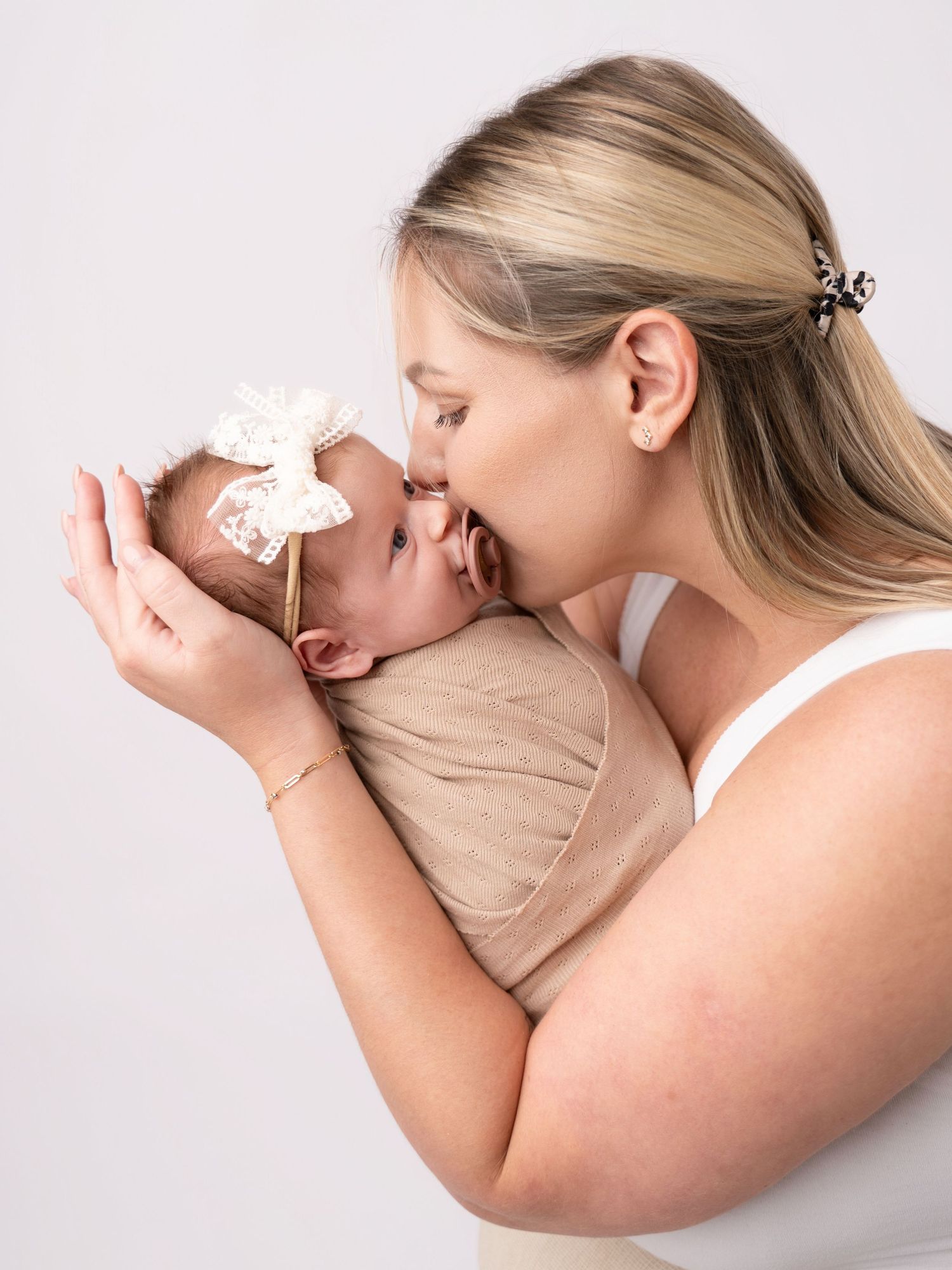 A mother tenderly kisses her newborn baby wrapped in a beige swaddle with a lace bow headband.