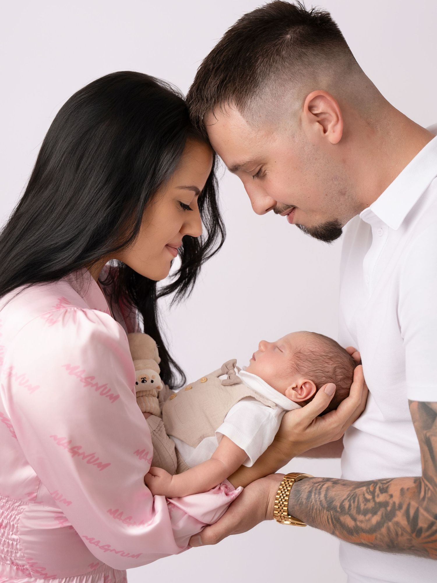 New parents tenderly gaze at their sleeping newborn baby, mother in pink silk robe, father with gold bracelet and tattoos.