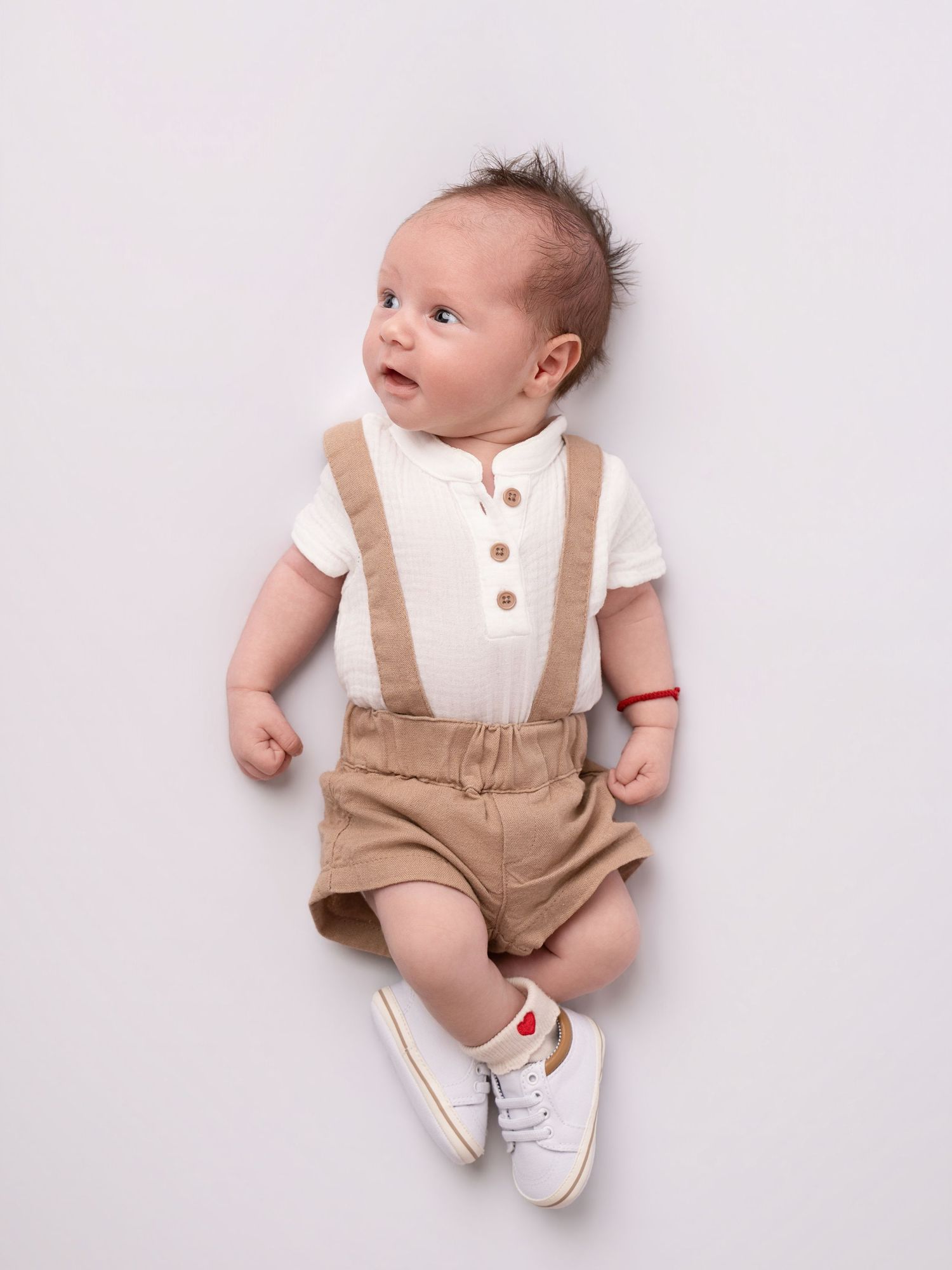 Adorable baby boy lying down wearing cream polo shirt with tan suspender shorts and white sneakers.