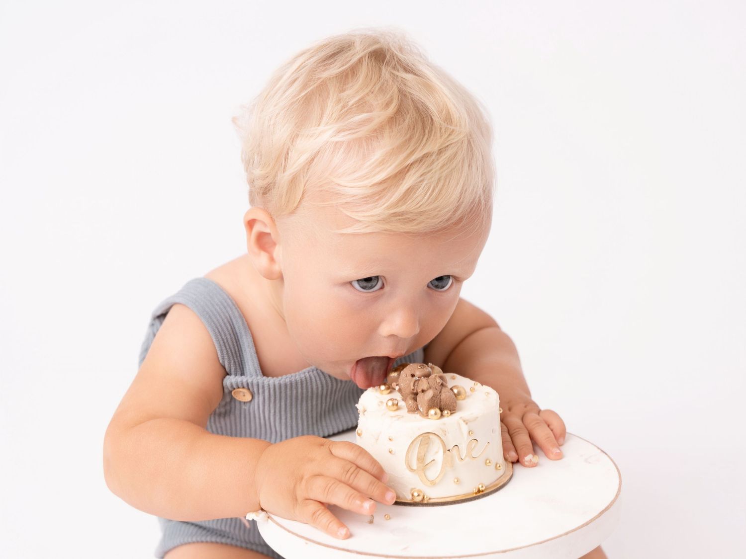 Adorable toddler in gray romper eating a small white birthday smash cake against a white background.