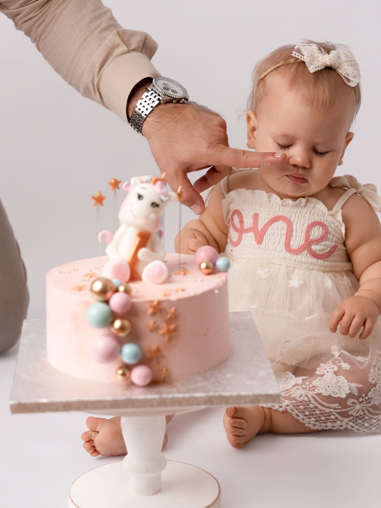 Baby girl in lace one birthday dress tastes cake frosting from fathers finger at cake smash session.