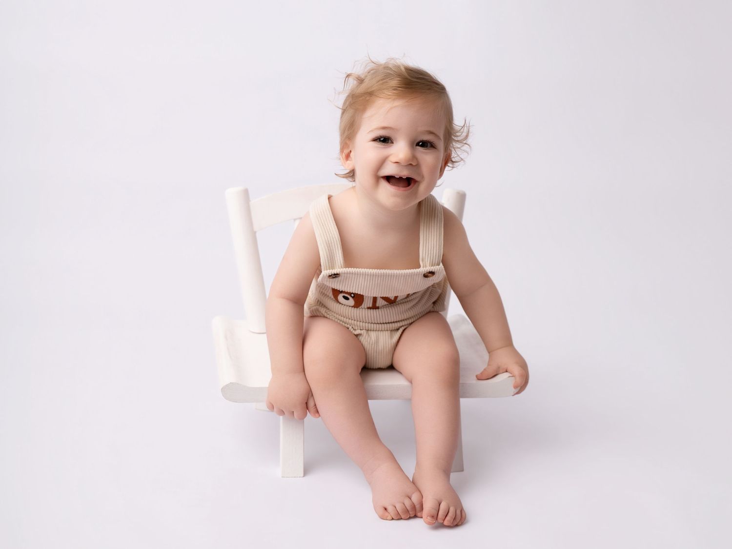 Happy laughing baby in beige romper sitting on small white chair against white background.