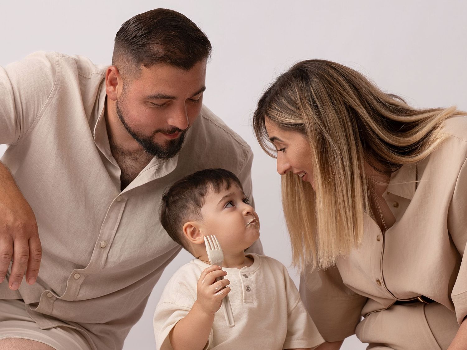A young family in matching beige outfits shares a tender moment with their toddler boy against a white background.