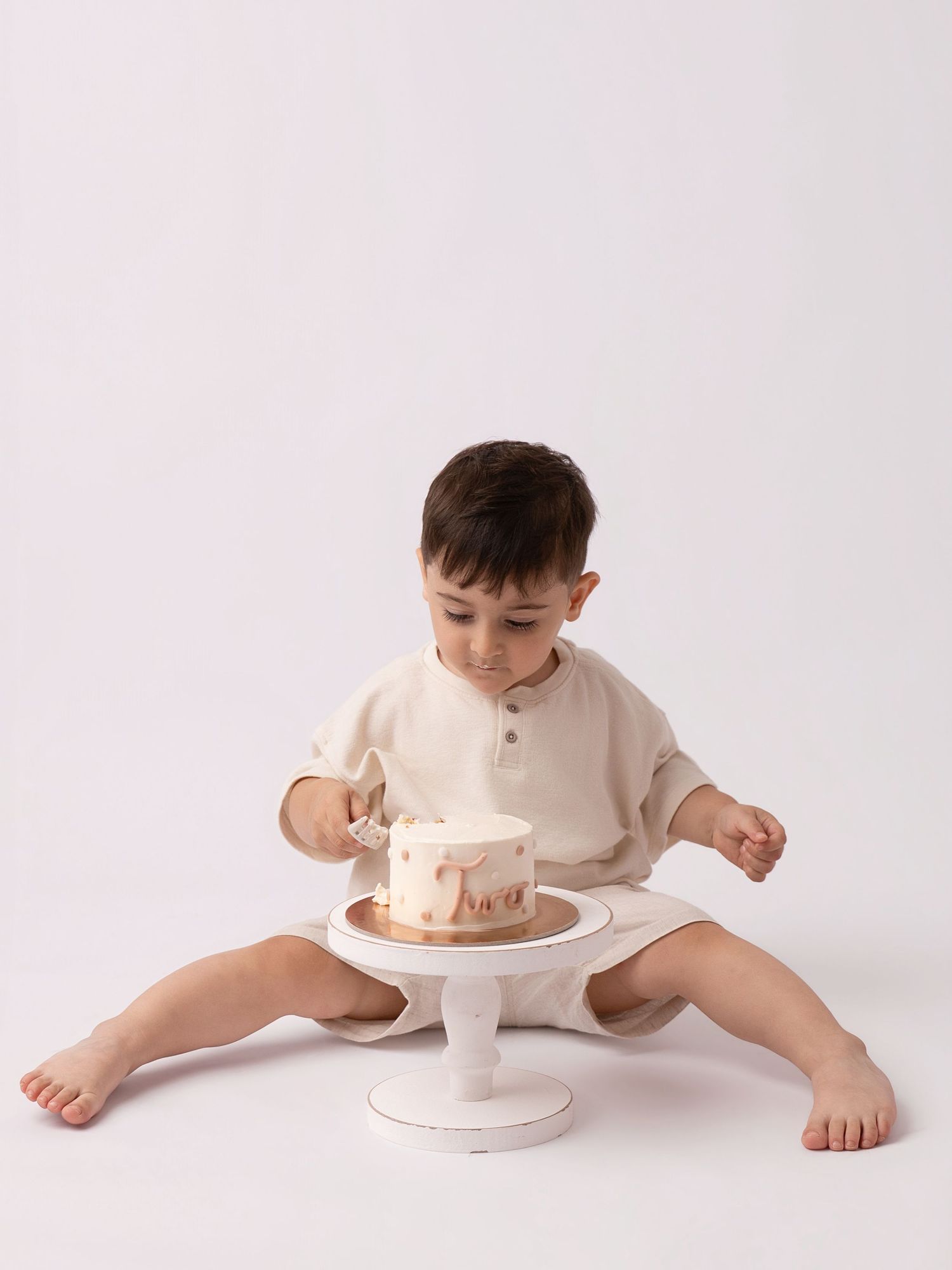 A toddler in a beige outfit sits on the floor exploring a small birthday cake on a white cake stand.