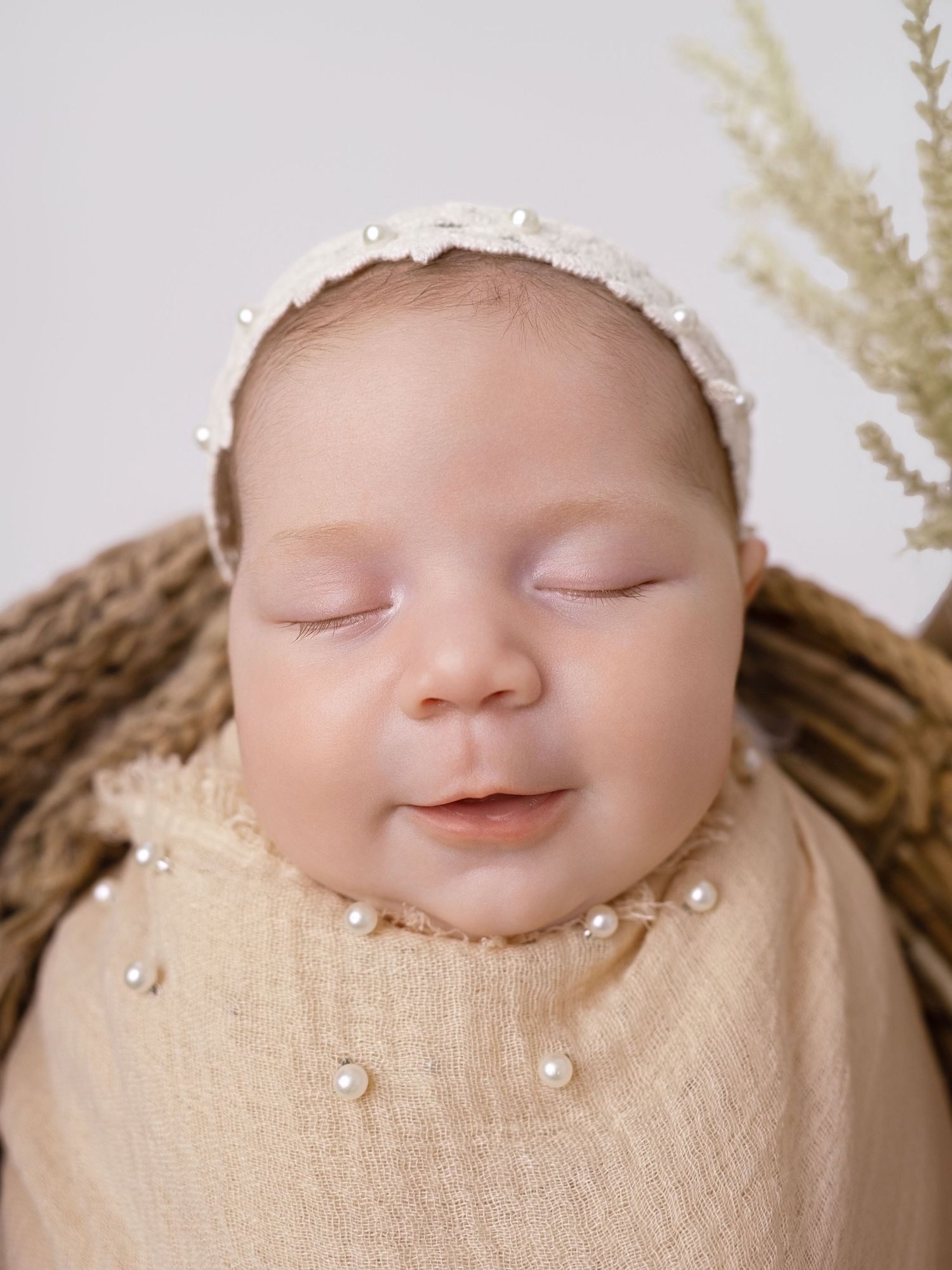 Sleeping newborn baby wrapped in cream fabric with pearl headband in a cozy basket, serene newborn photography.