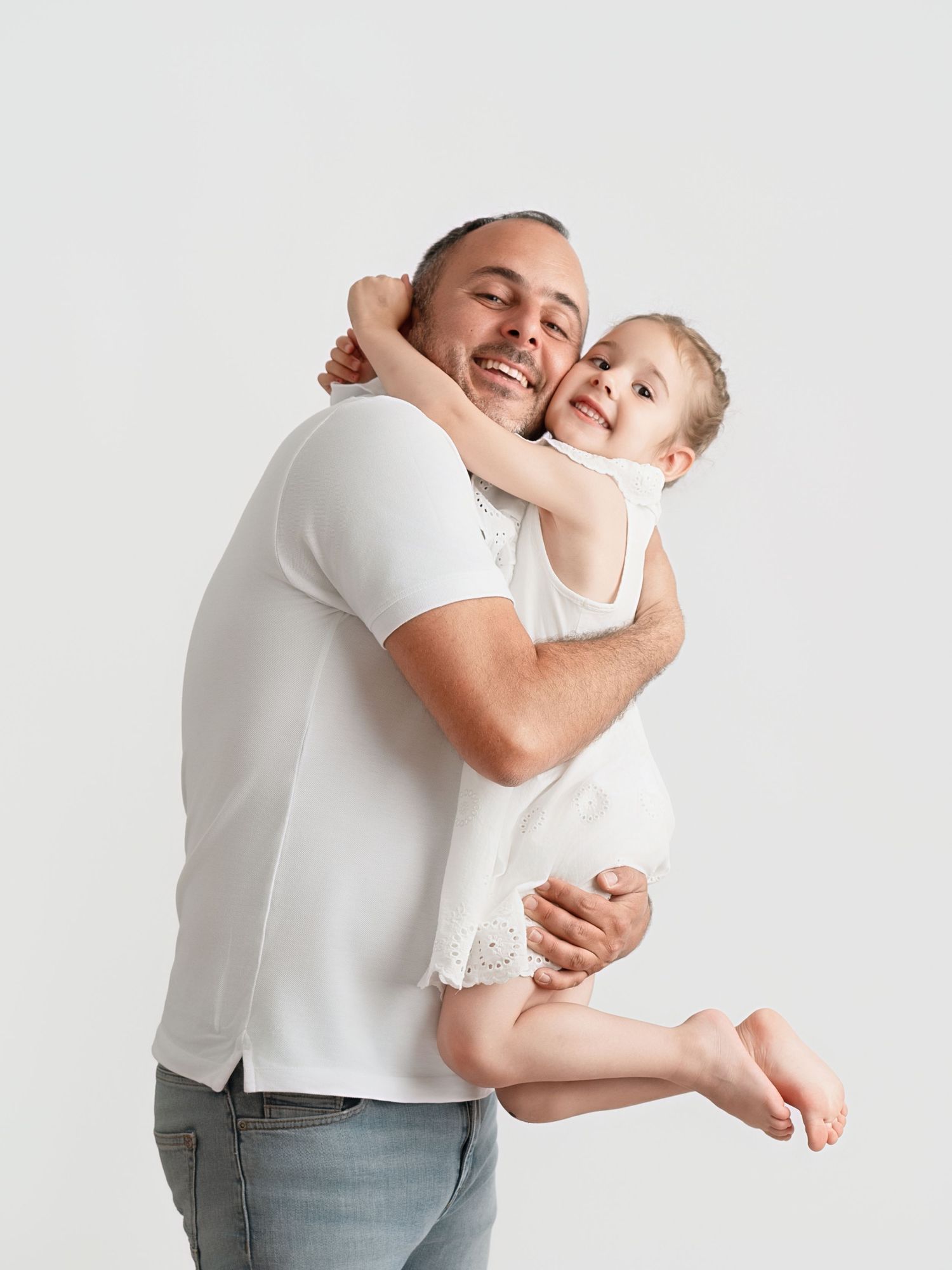 A smiling man in a white t-shirt holds a happy young girl in a white dress against a light background.