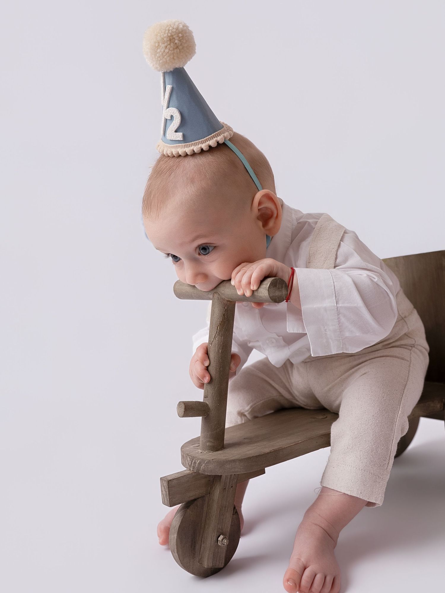 Baby wearing a blue party hat with pom-pom sitting on a wooden toy scooter, celebrating a half birthday.