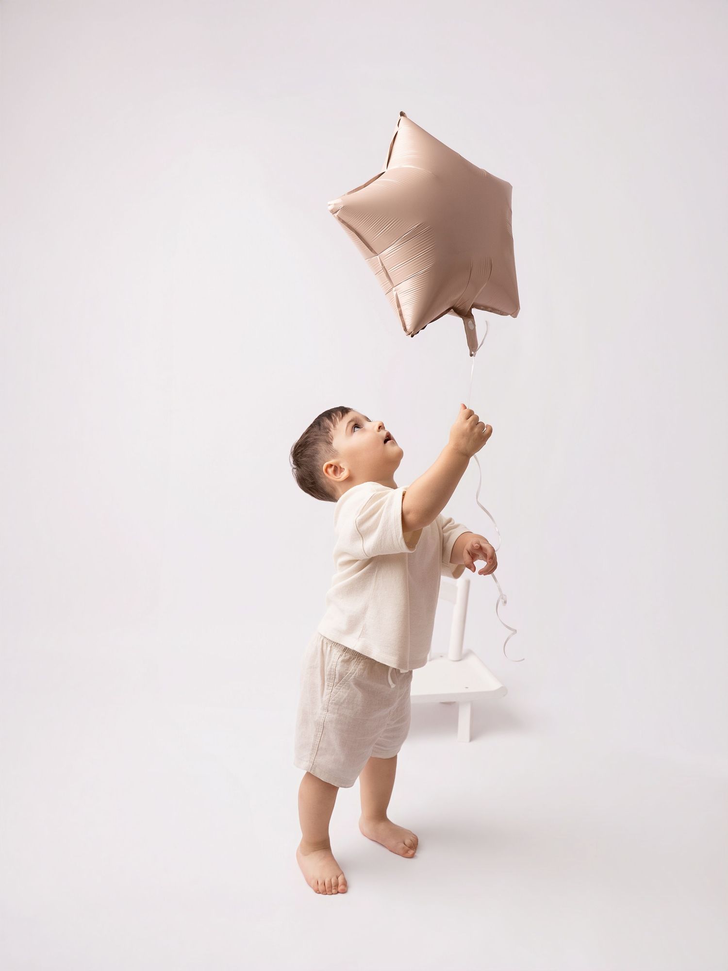 Cute toddler boy reaching up to hold a rose gold star balloon in a white studio setting.