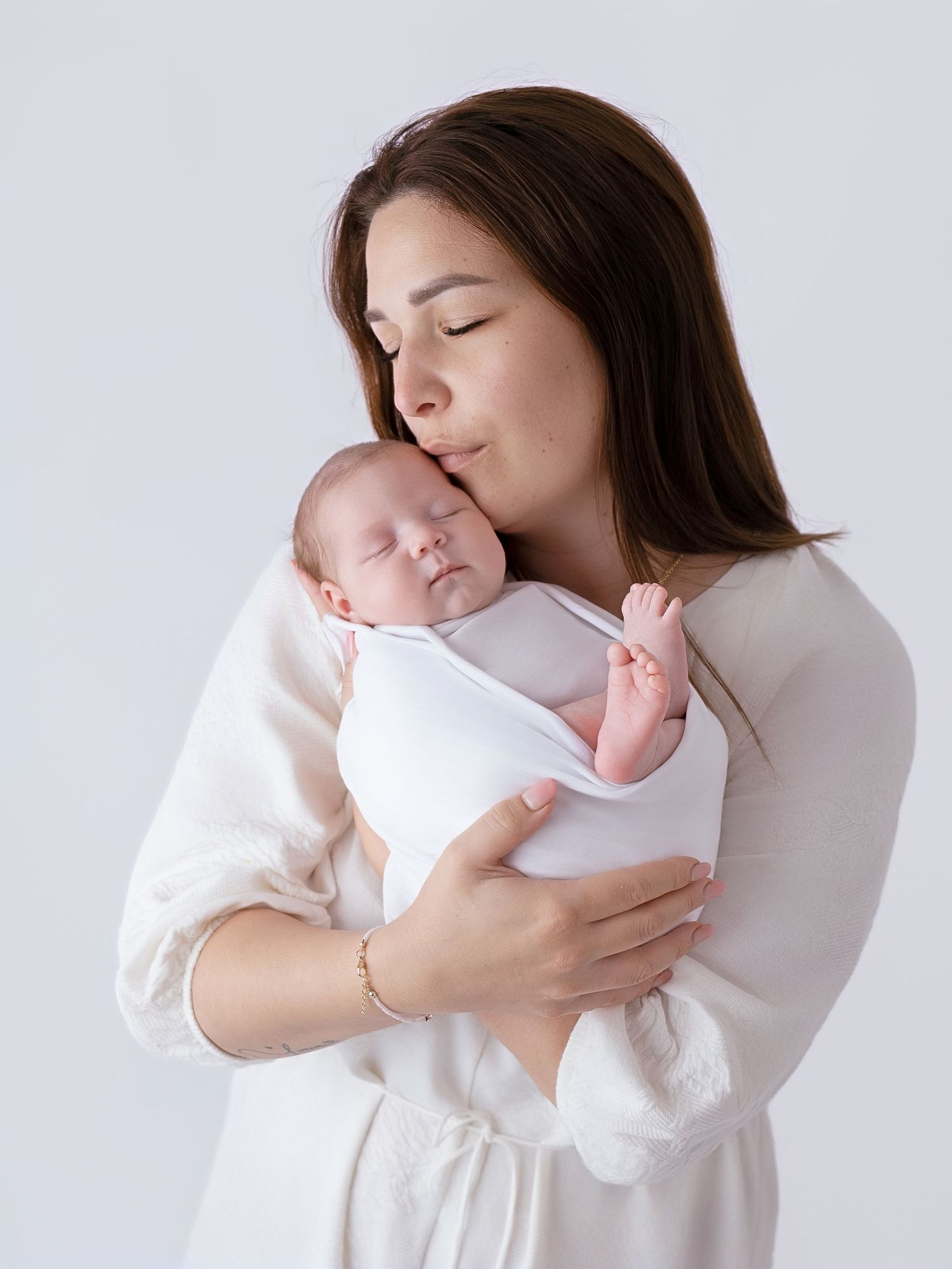 Loving mother with dark hair gently kisses sleeping newborn baby wrapped in white swaddle cloth.