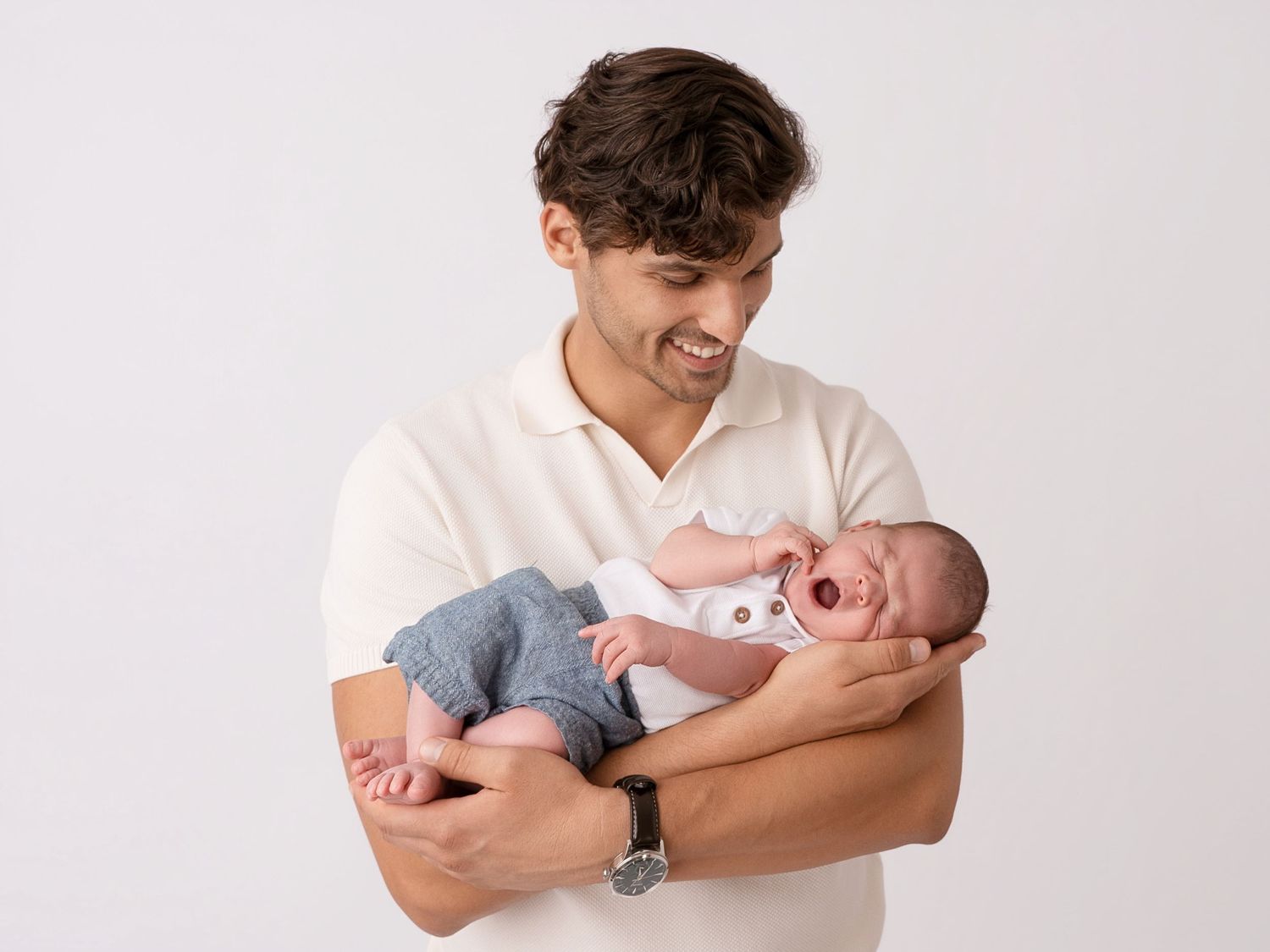 Young man in white polo shirt smiling while cradling a sleeping newborn baby in his arms.