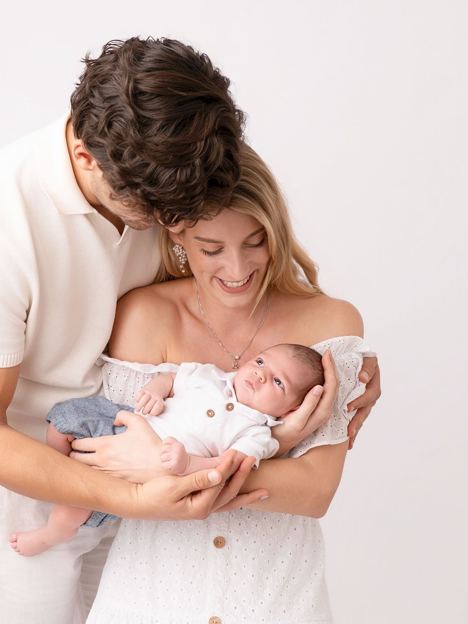 Happy couple in white outfits cradling their newborn baby during a professional studio photography session.