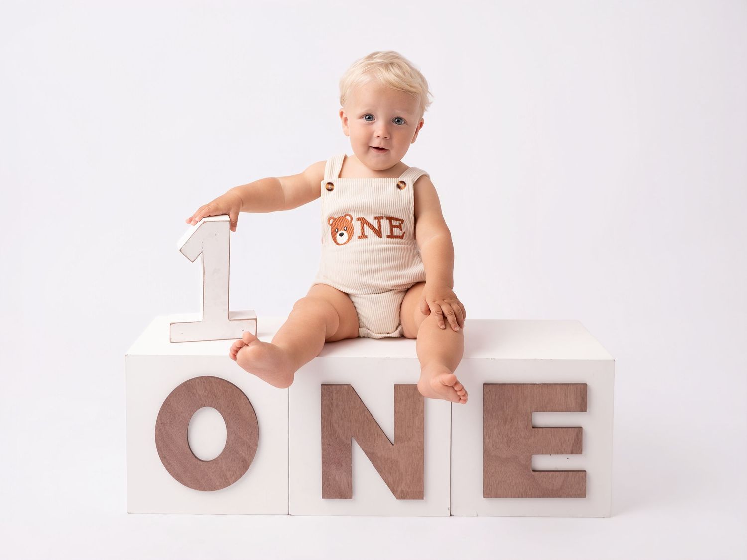Baby's first birthday photo shoot with ONE letter blocks, wearing a bear romper, sitting on white cube.