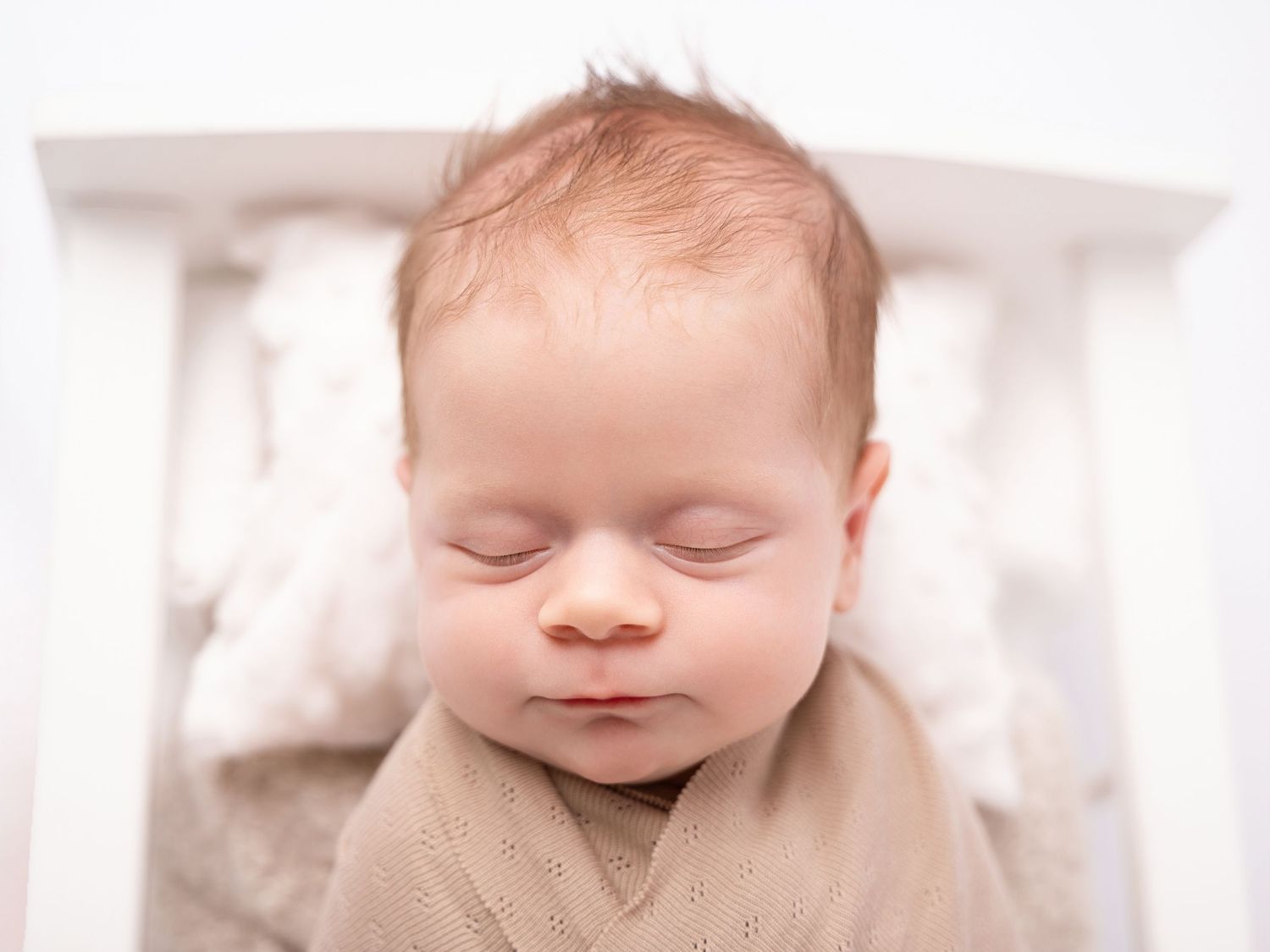 Peaceful newborn baby sleeping swaddled in a beige wrap inside a white crib during a professional newborn photo session.