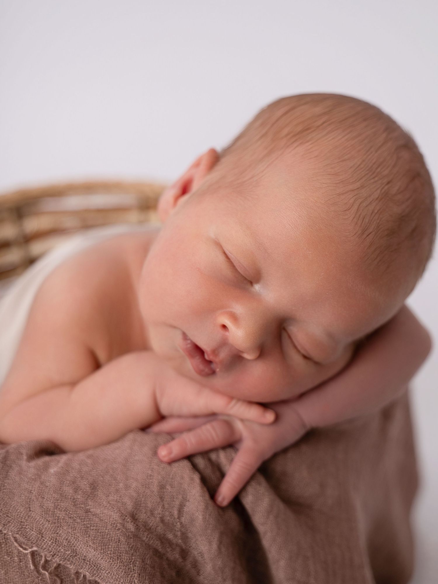 Peaceful newborn baby sleeping curled up in a wicker basket with a soft brown blanket in a studio portrait.