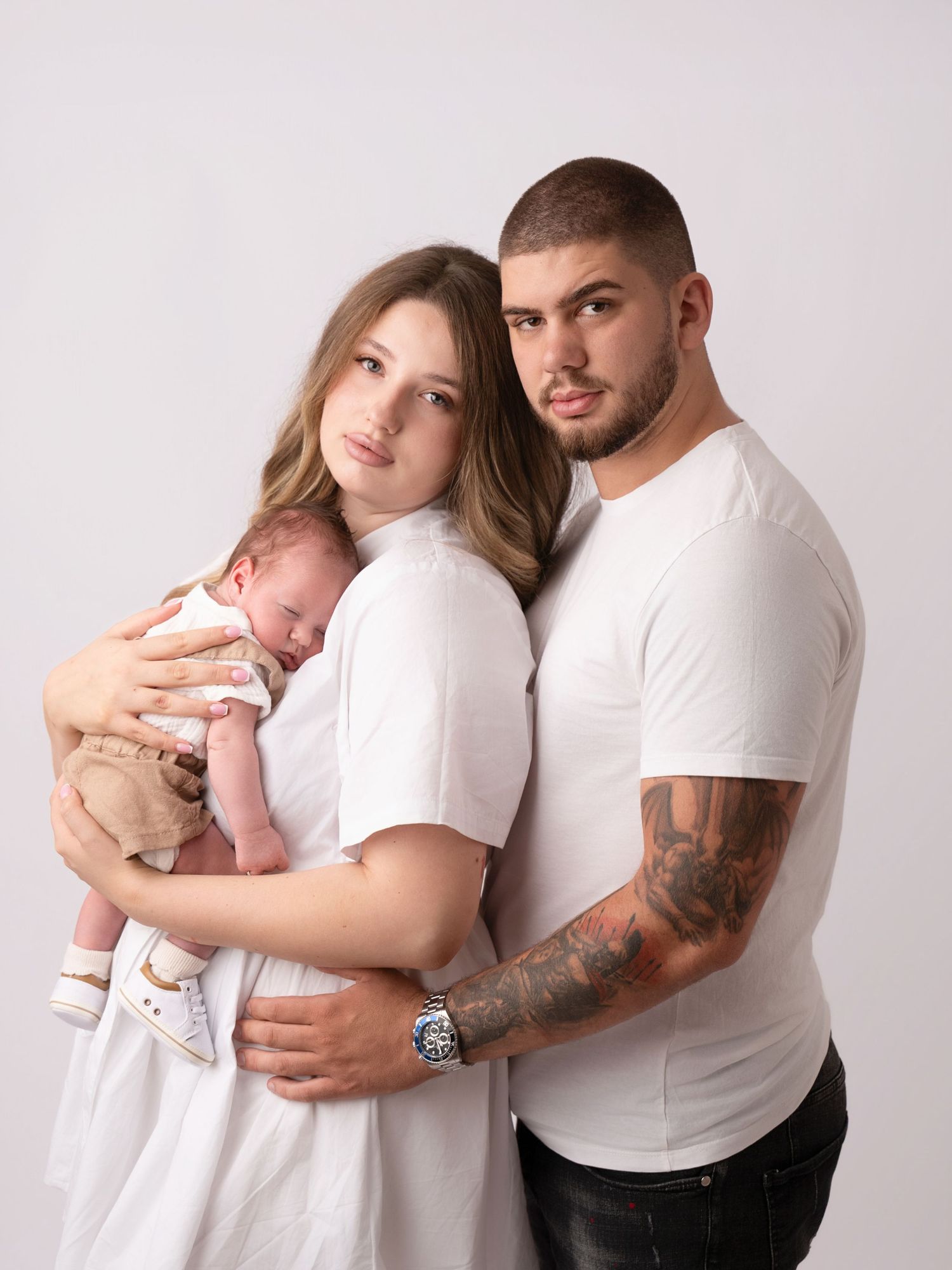 Young couple in white shirts posing with their newborn baby in a professional studio portrait photo.